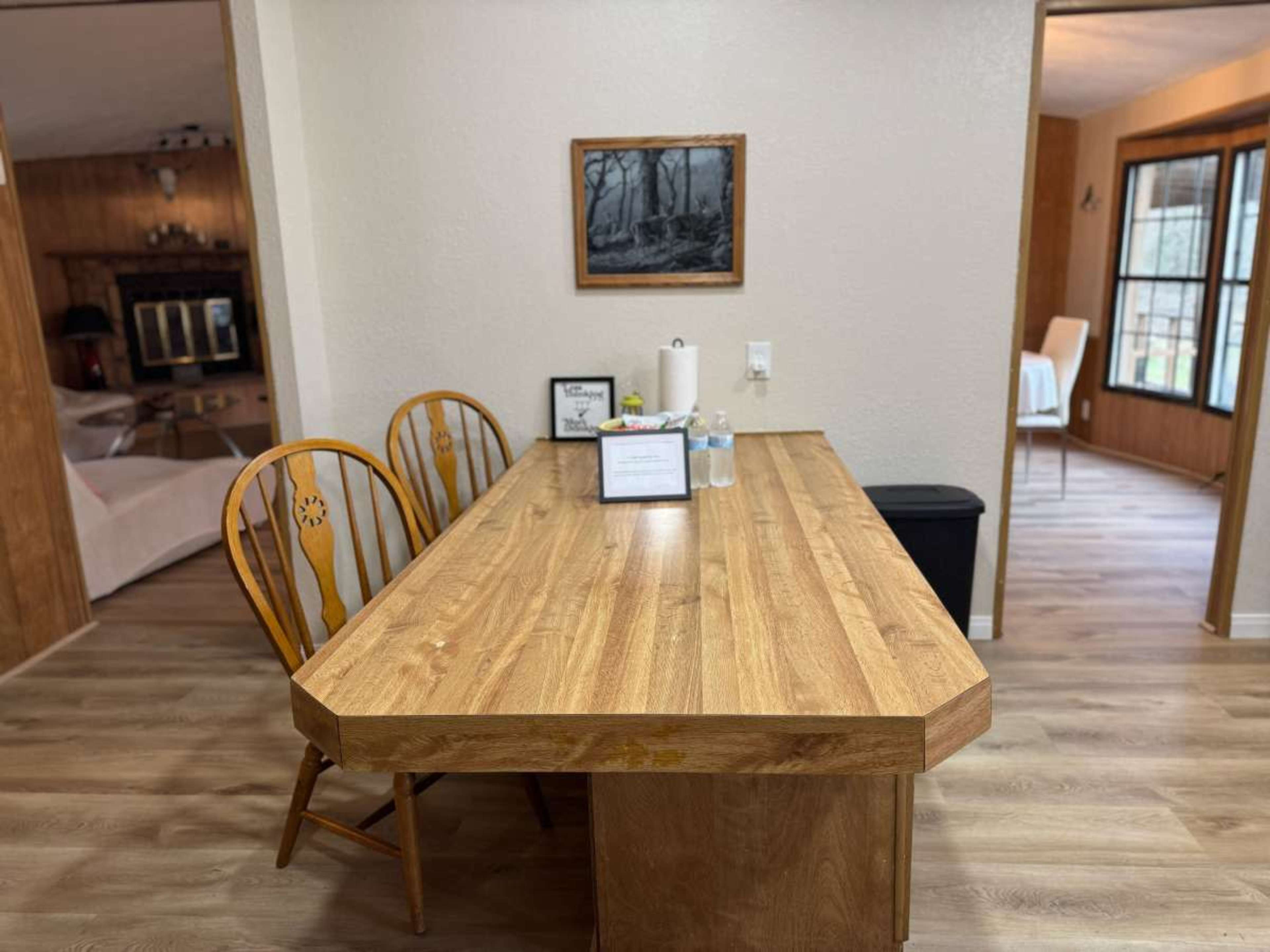 A wooden dining table with two chairs is positioned in a well-lit room, with a water bottle and a sign on the table, and a view of another room through an open doorway.