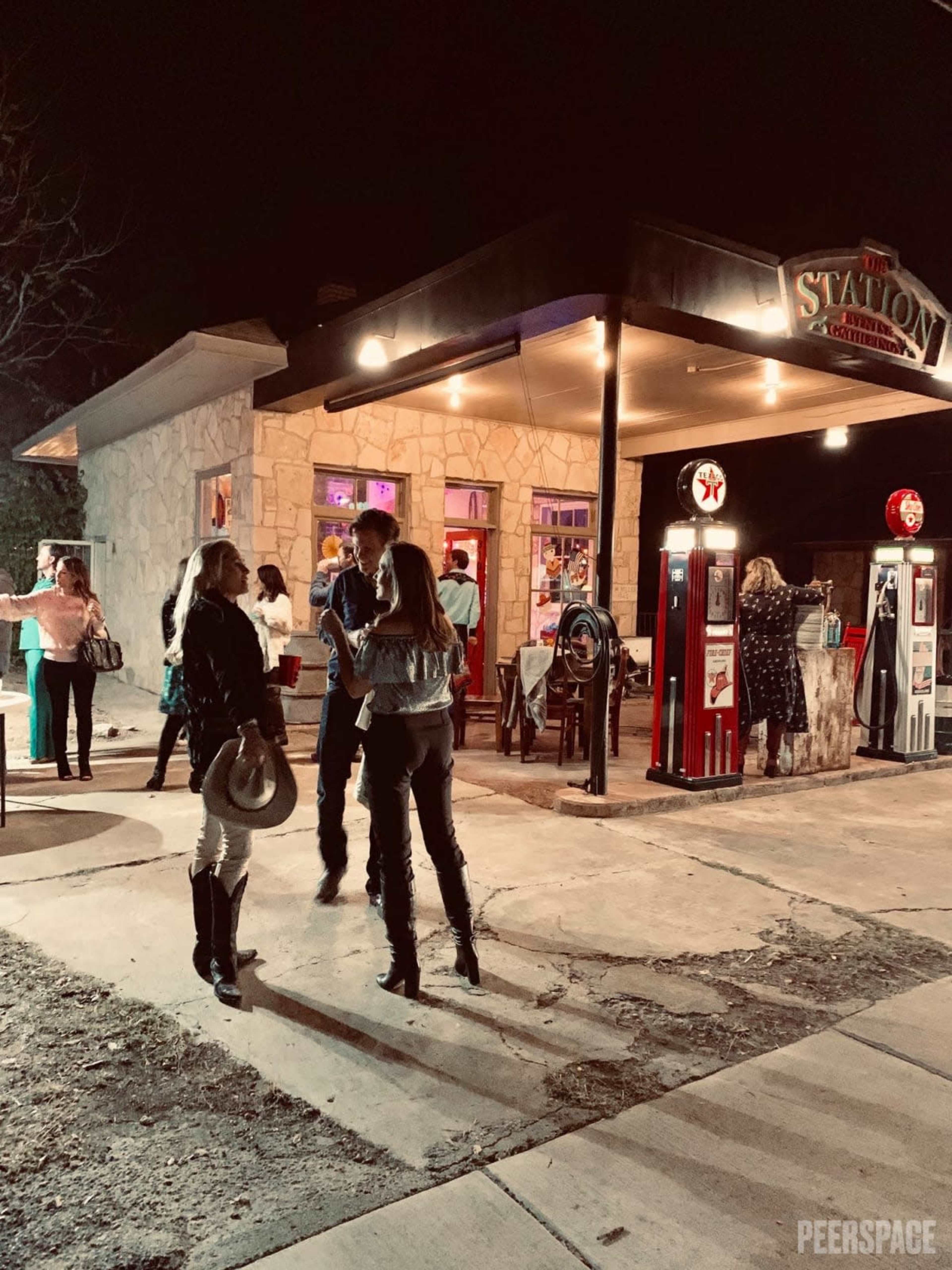 A group of people mingles outside a stone building with neon lights and vintage gas pumps on a busy street at night.