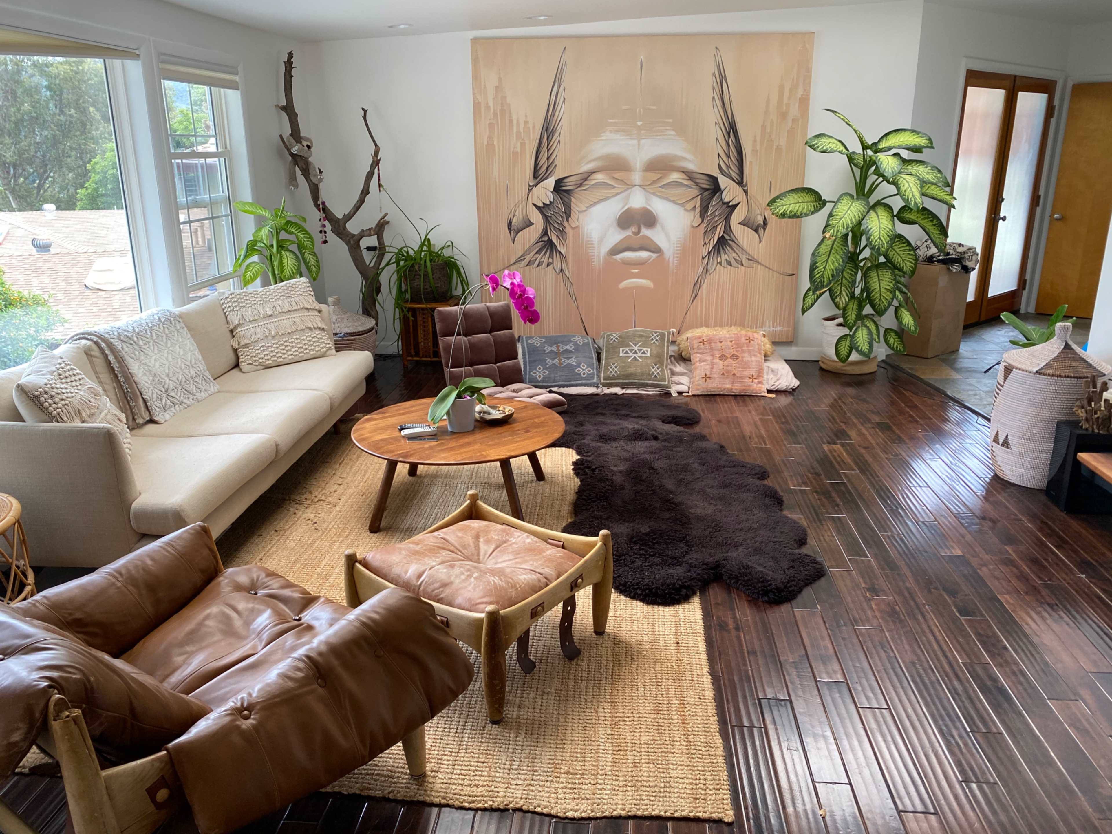 The living room features a large mural on the wall, a circular wooden coffee table surrounded by various seating arrangements, and a mix of decorative plants.