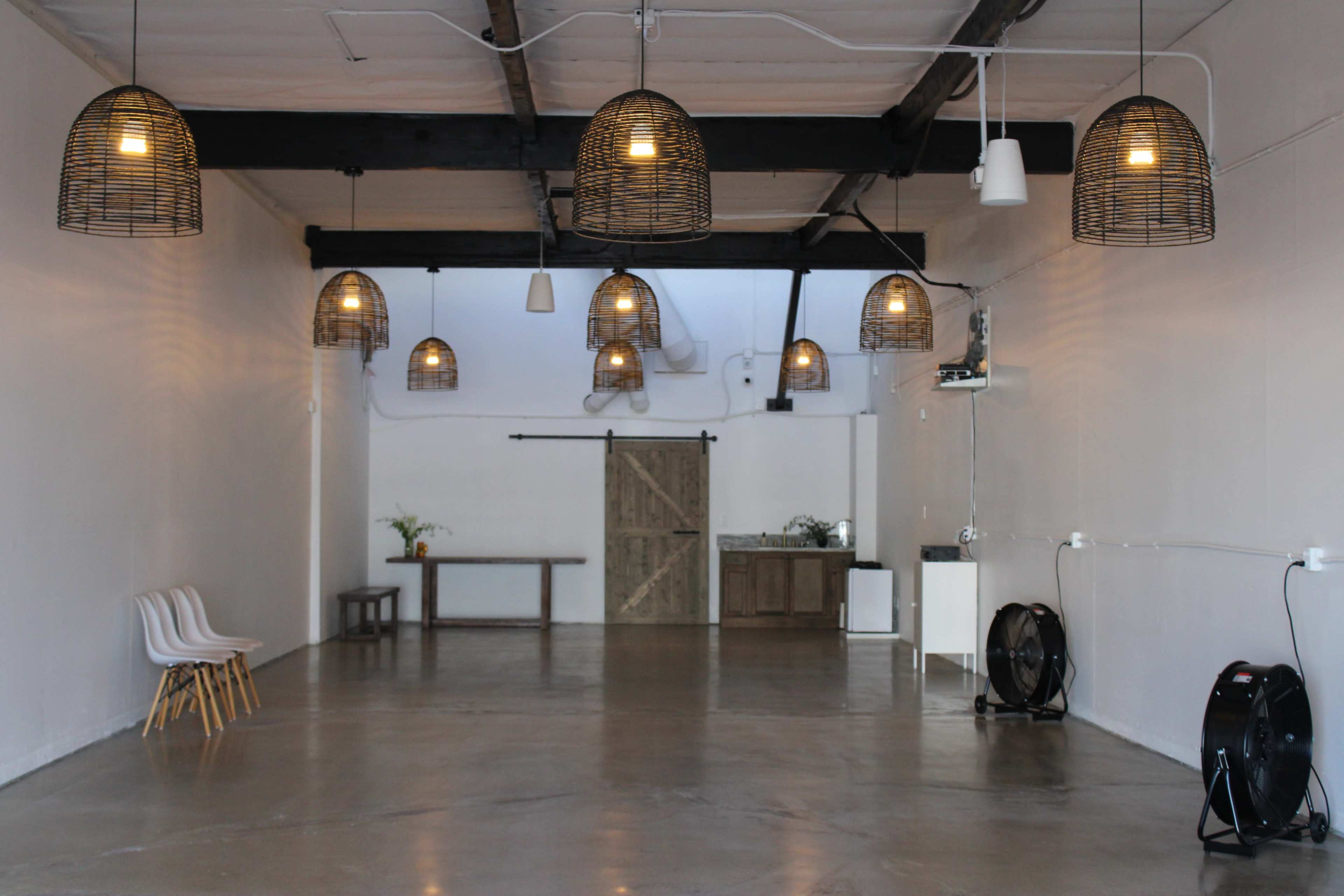 The image shows a spacious, empty room with concrete flooring, illuminated by pendant lights in woven baskets, featuring a wooden table, white chairs, and a sliding barn door.