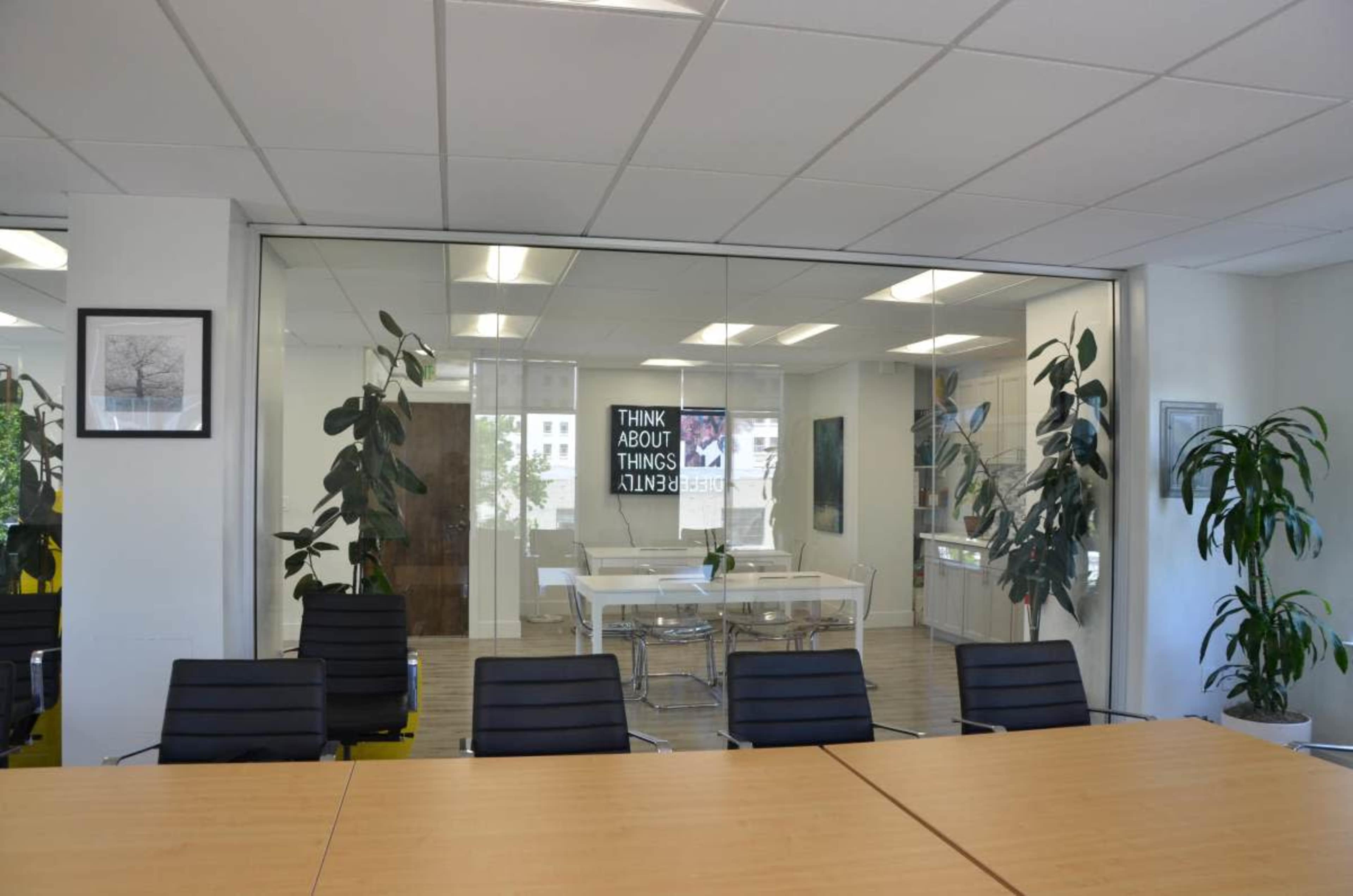 A modern office space features a glass-walled conference room with a wooden table and black chairs, surrounded by greenery and accented with motivational wall art.