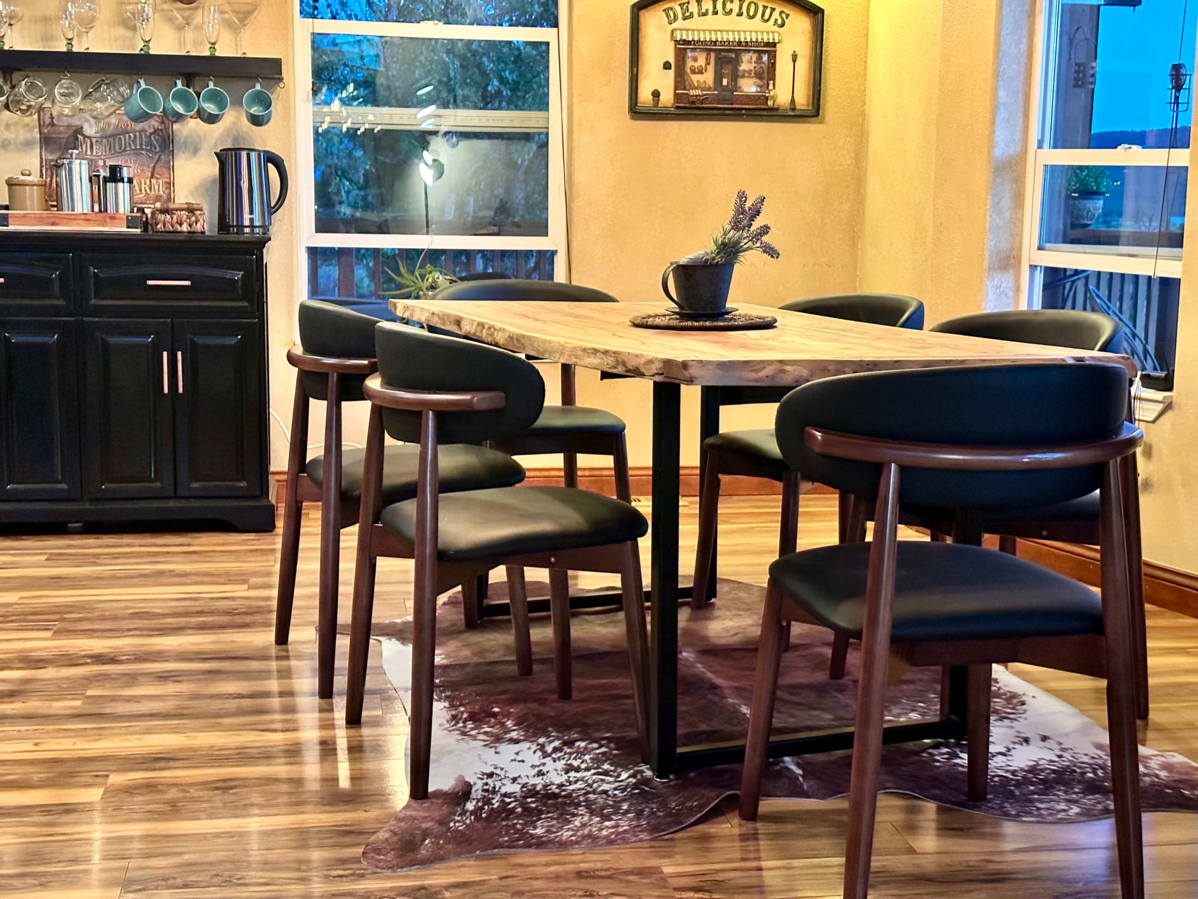 The image shows a dining area with a wooden table surrounded by six chairs, set against a backdrop of large windows and a kitchen cabinet.