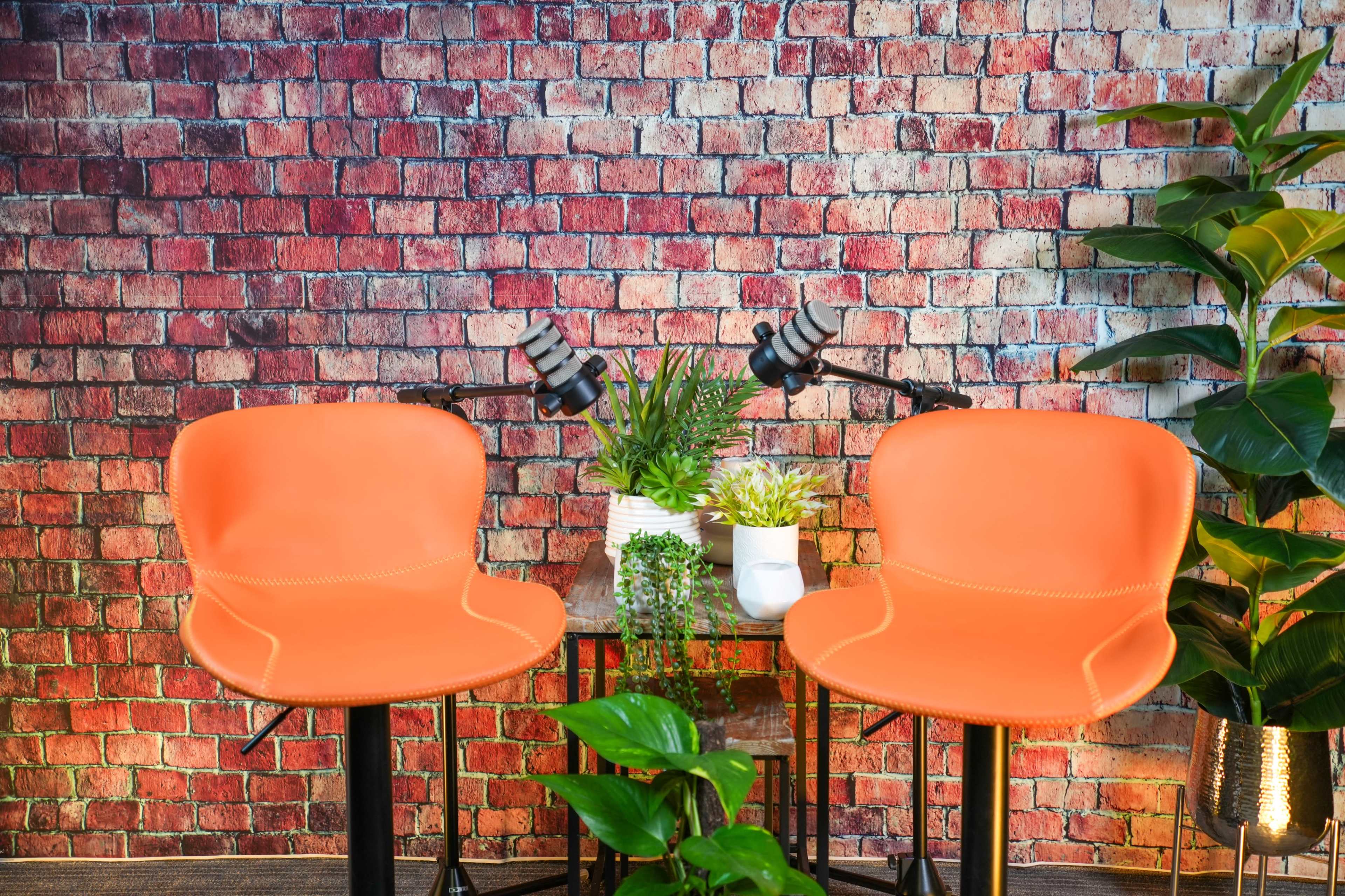 Two orange chairs are positioned at a small table surrounded by various plants and a brick wall backdrop.