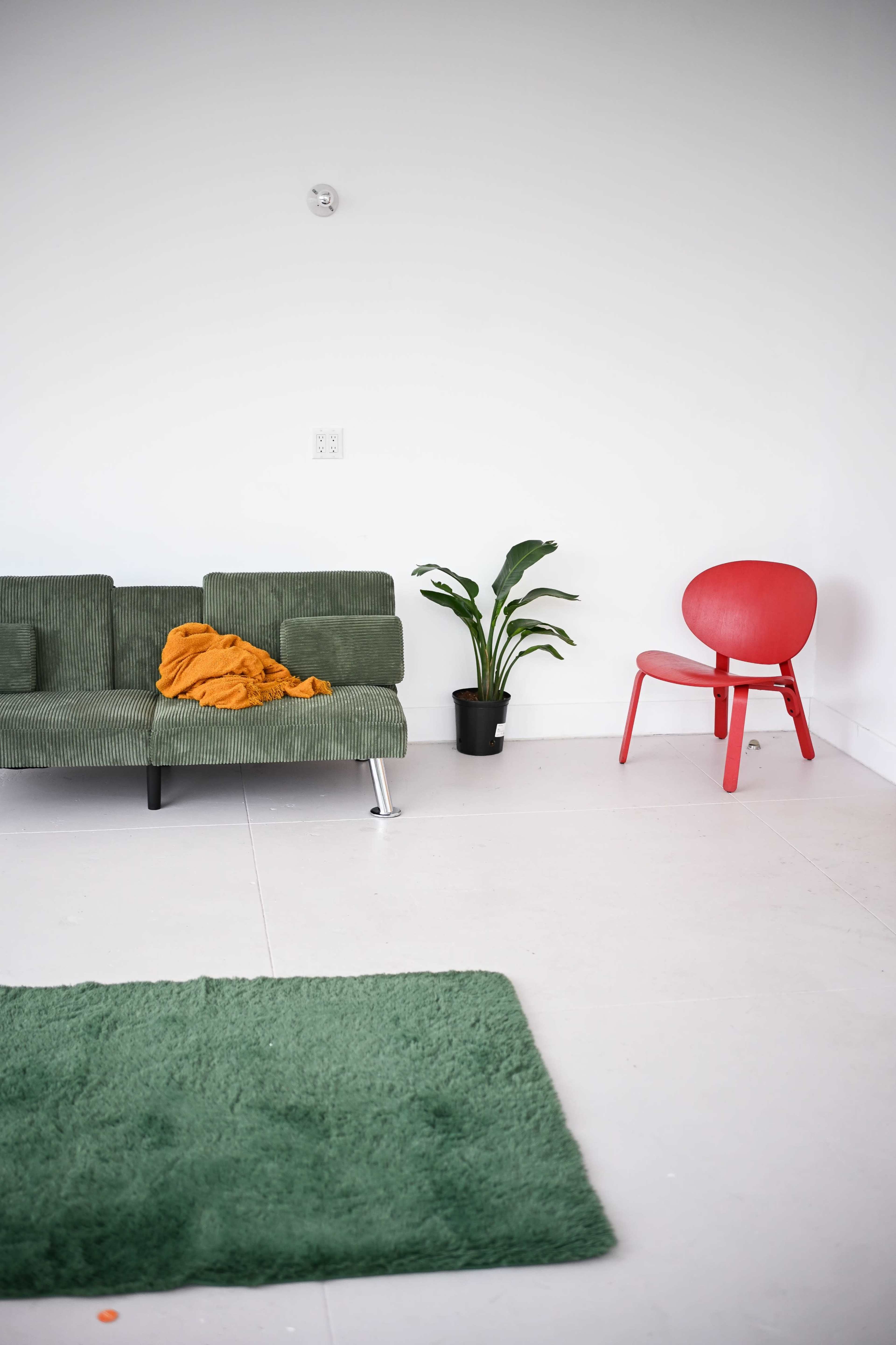 A green sofa with an orange blanket sits beside a potted plant and a red chair in a minimalist room.