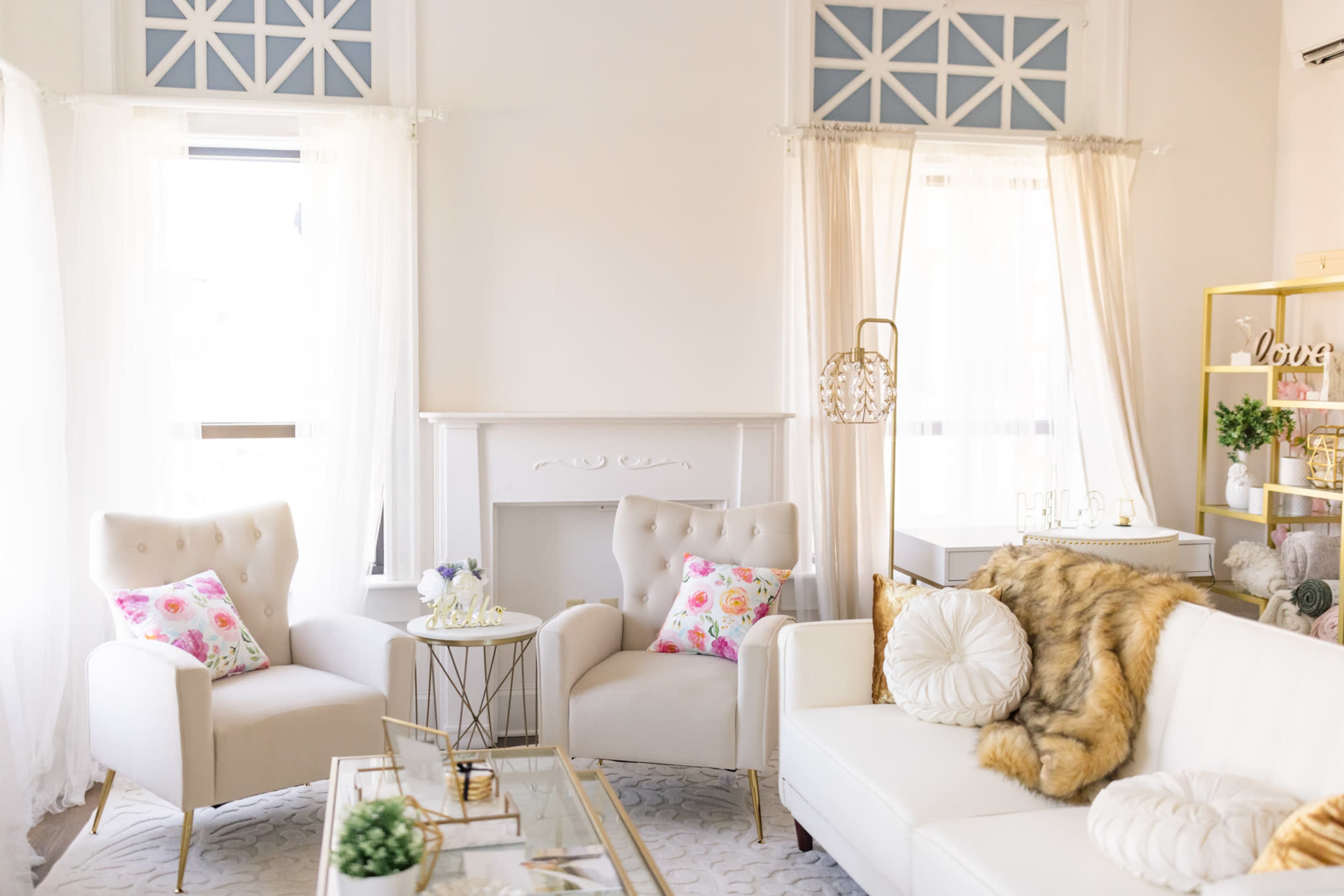 A bright and airy living room furnished with two light-colored armchairs, a white sofa, and a coffee table, all surrounded by large windows with sheer curtains.