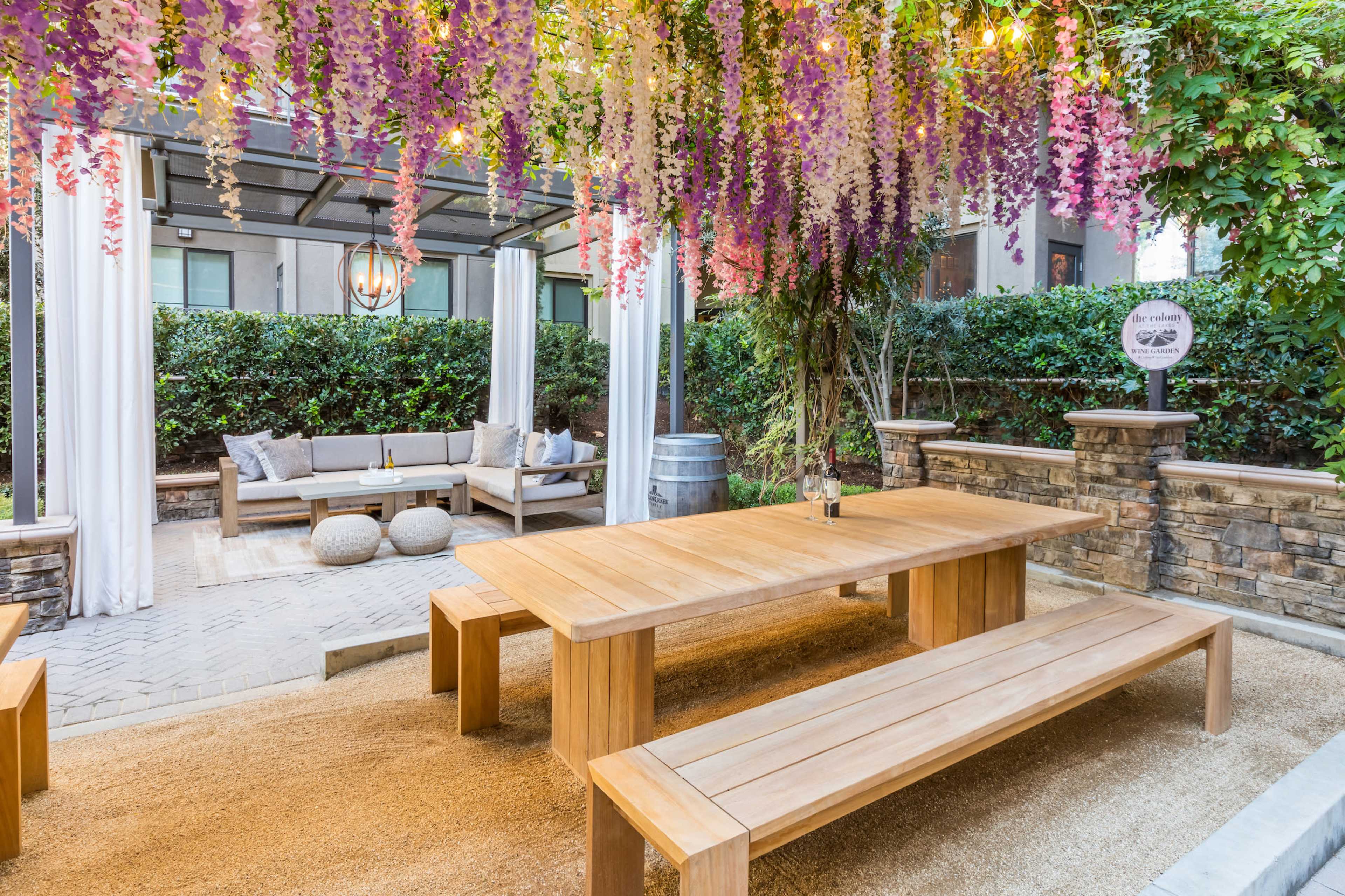 A wooden dining table with matching benches is situated under a canopy of hanging flowers and lights in an outdoor seating area.