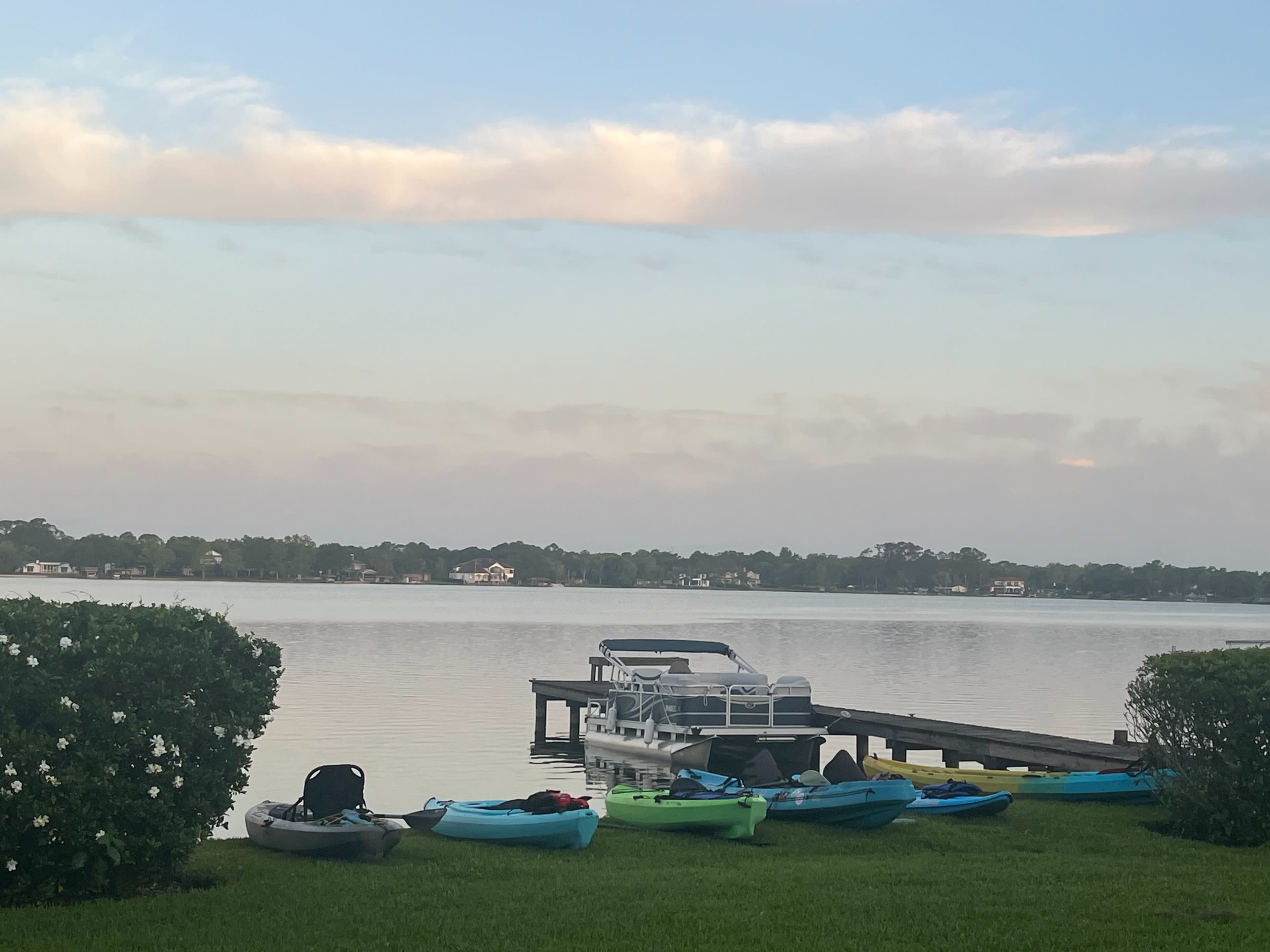Kayaks are lined up on the grassy shore next to a calm lake, with a wooden dock extending into the water.