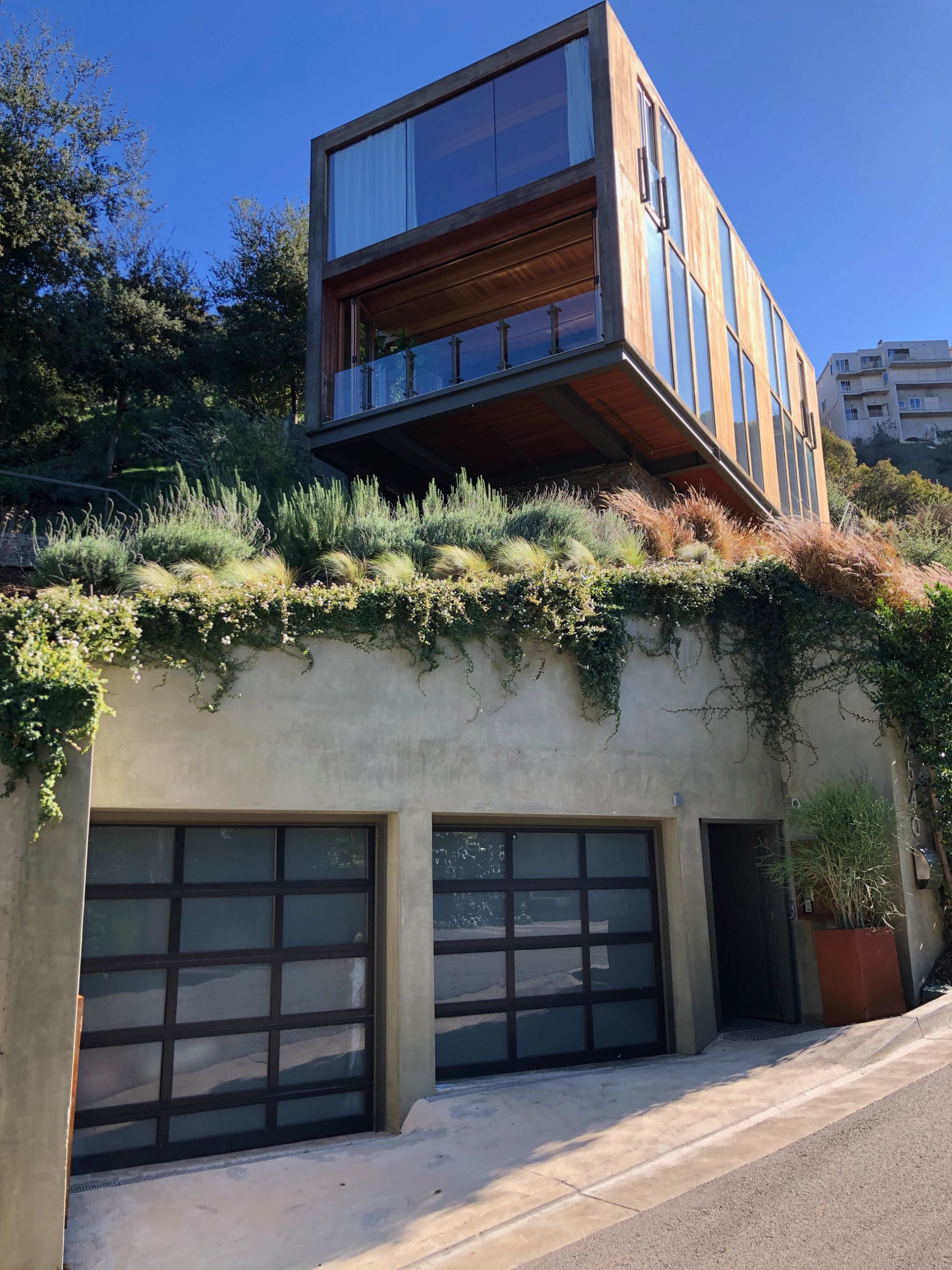 A modern house with a cantilevered upper section overlooks a garage, surrounded by greenery and situated on a slope.