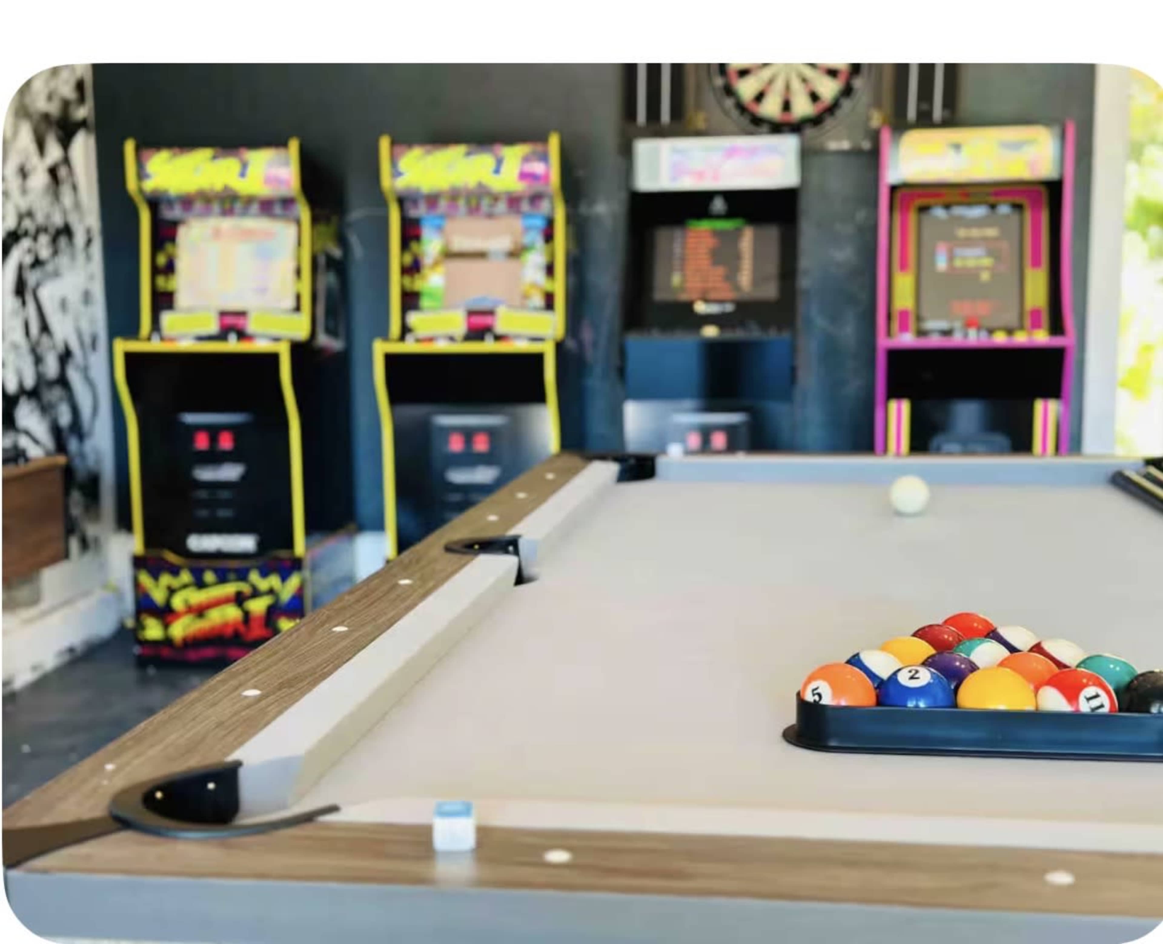 The image shows a billiards table with a rack of colored pool balls in the foreground and several vintage arcade games in the background.