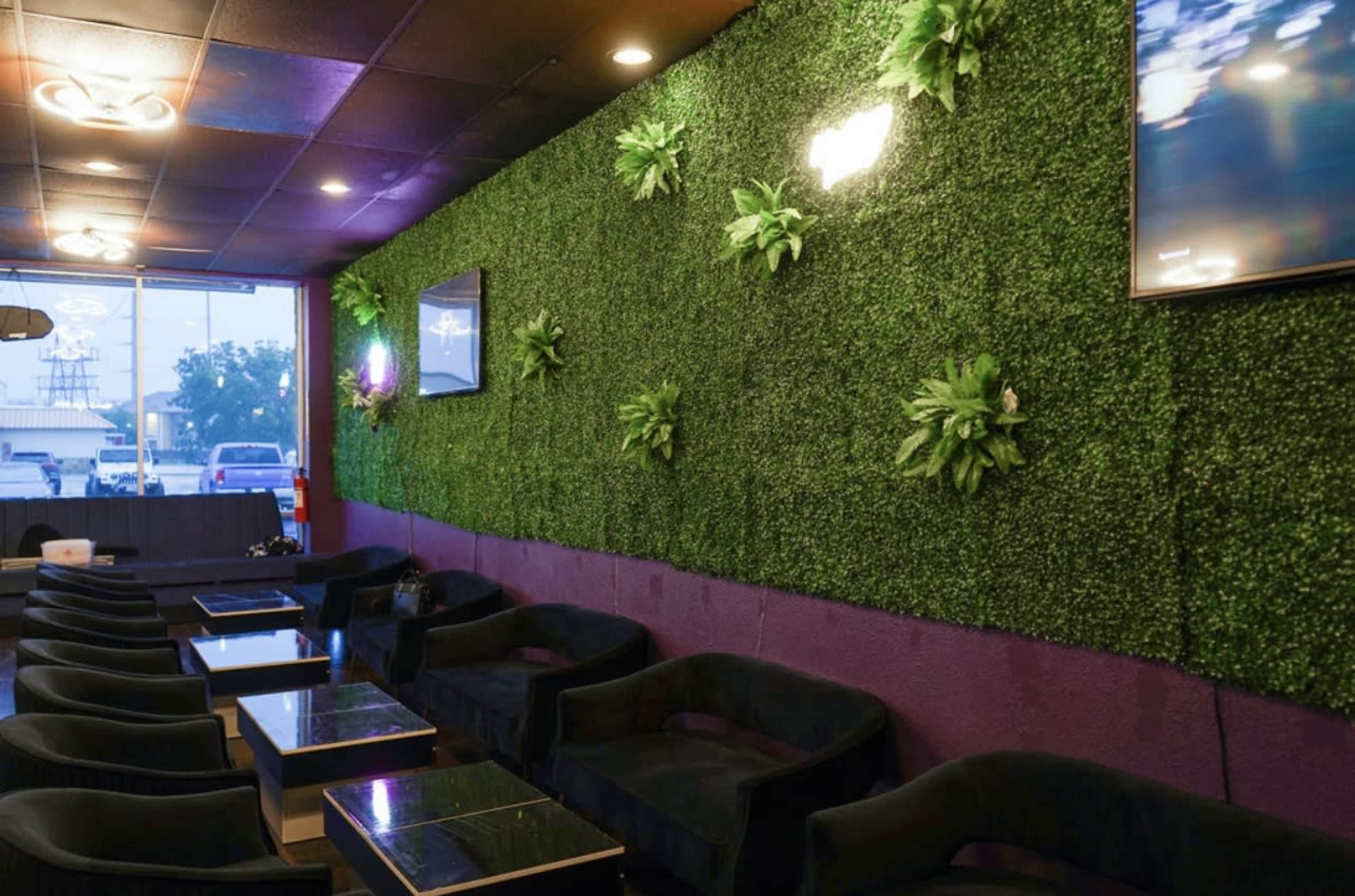 A lounge area with black sofas and a green wall adorned with artificial plants and two television screens.
