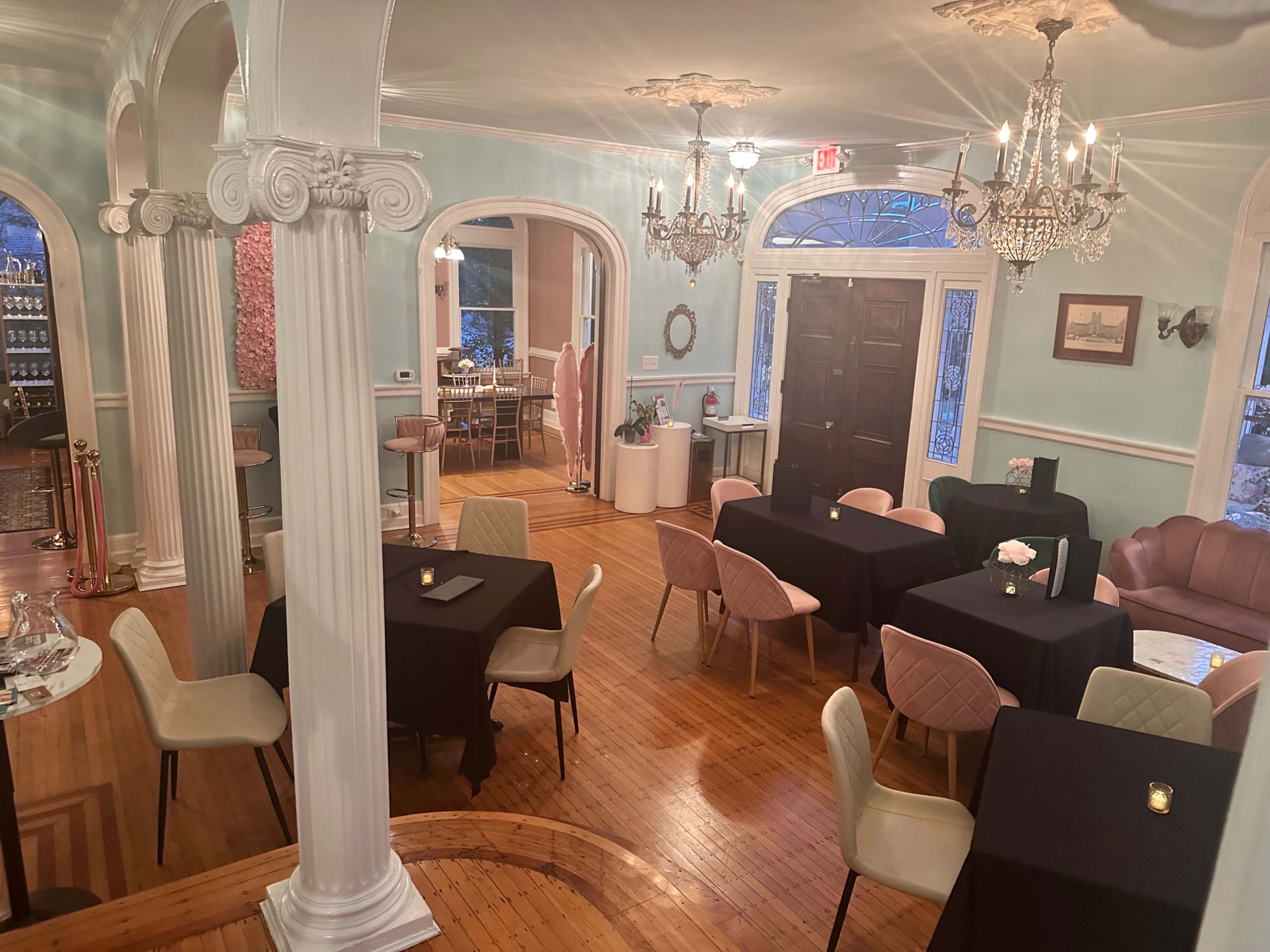 A dining area with several tables set for guests, featuring pale blue walls and decorative columns, illuminated by chandeliers.