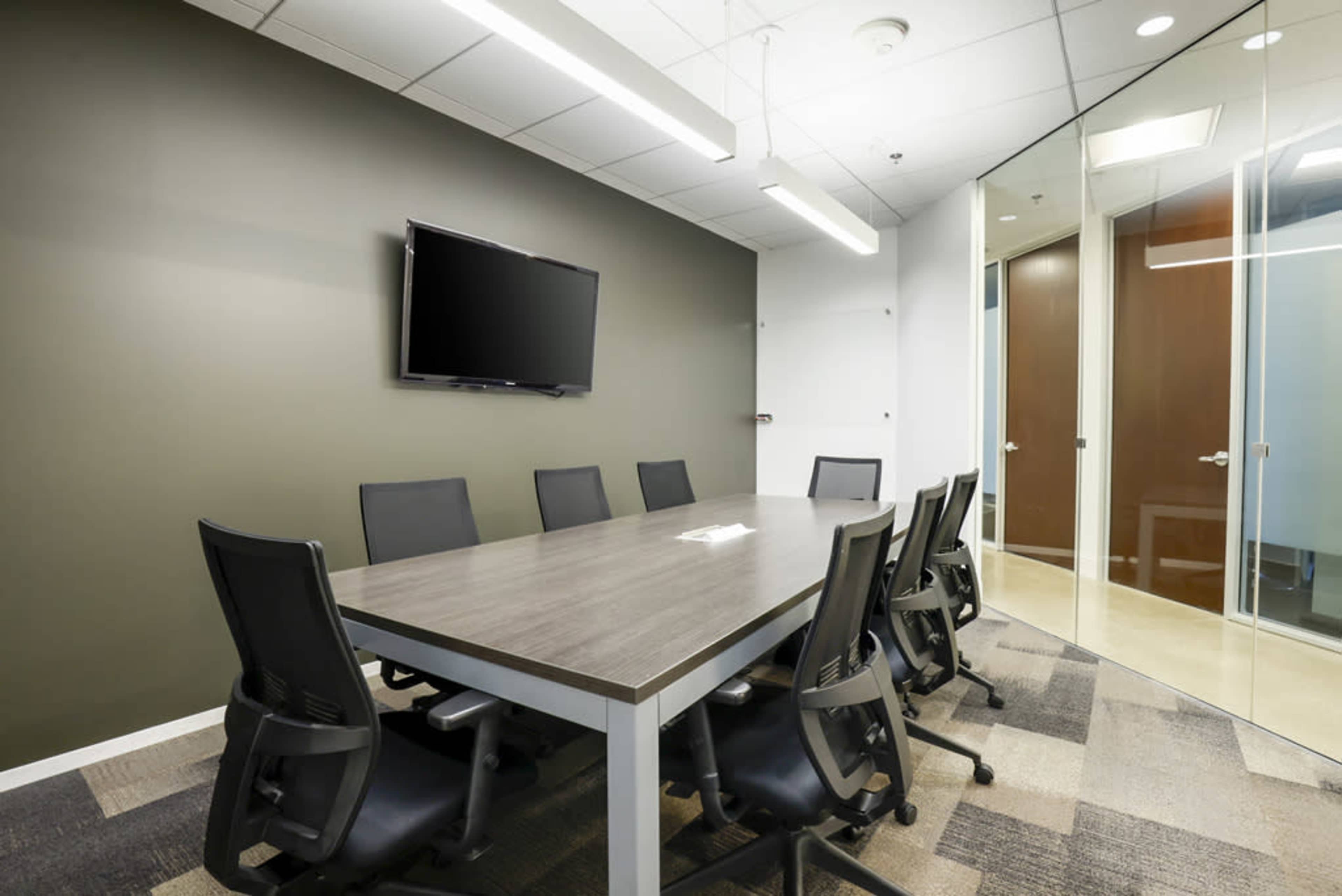 A modern conference room features a long table surrounded by black office chairs, with a wall-mounted television and glass-paneled doors.