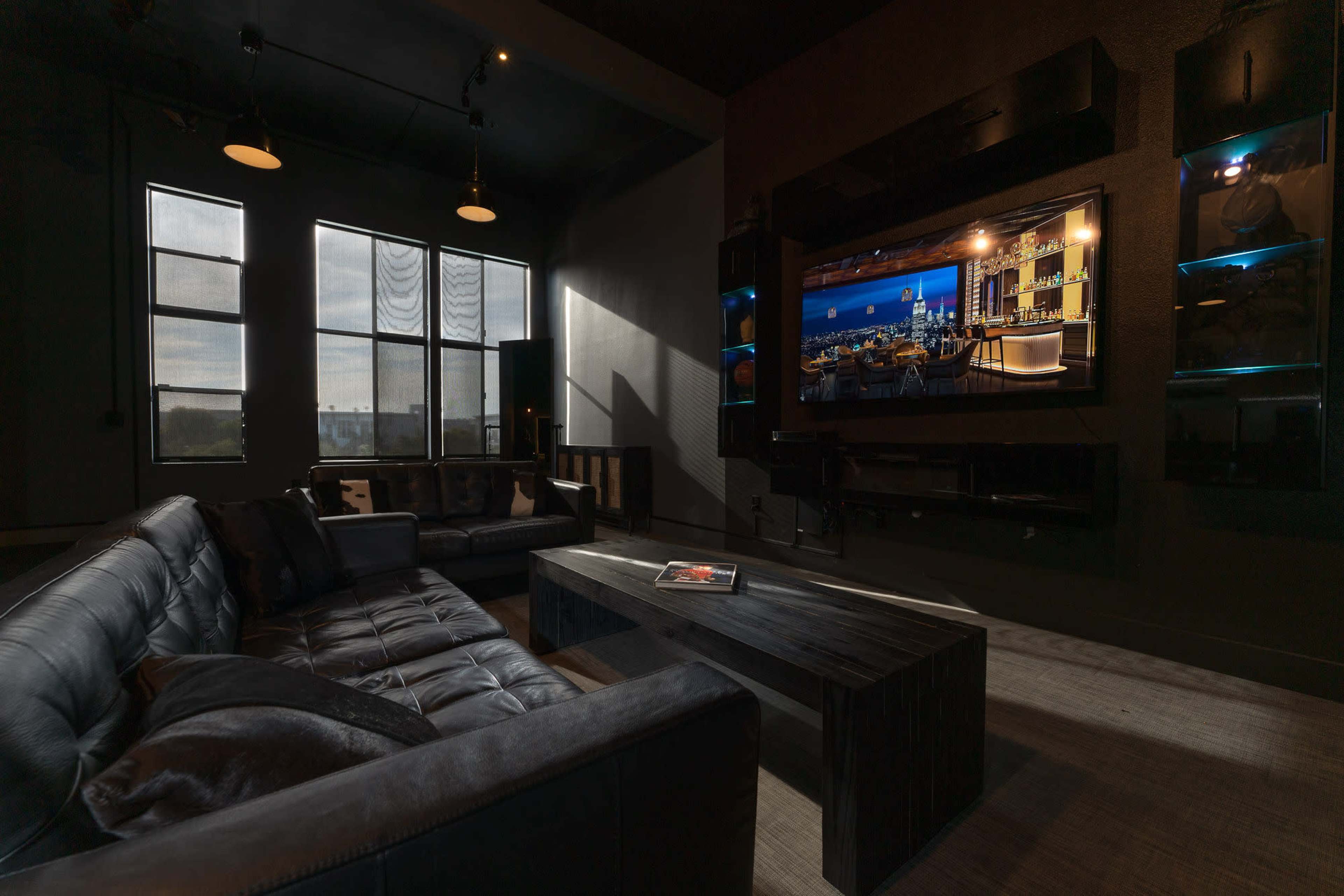 A modern living room with a black leather sofa, a wooden coffee table, and a large flat-screen TV mounted on the wall, illuminated by natural light from the windows.