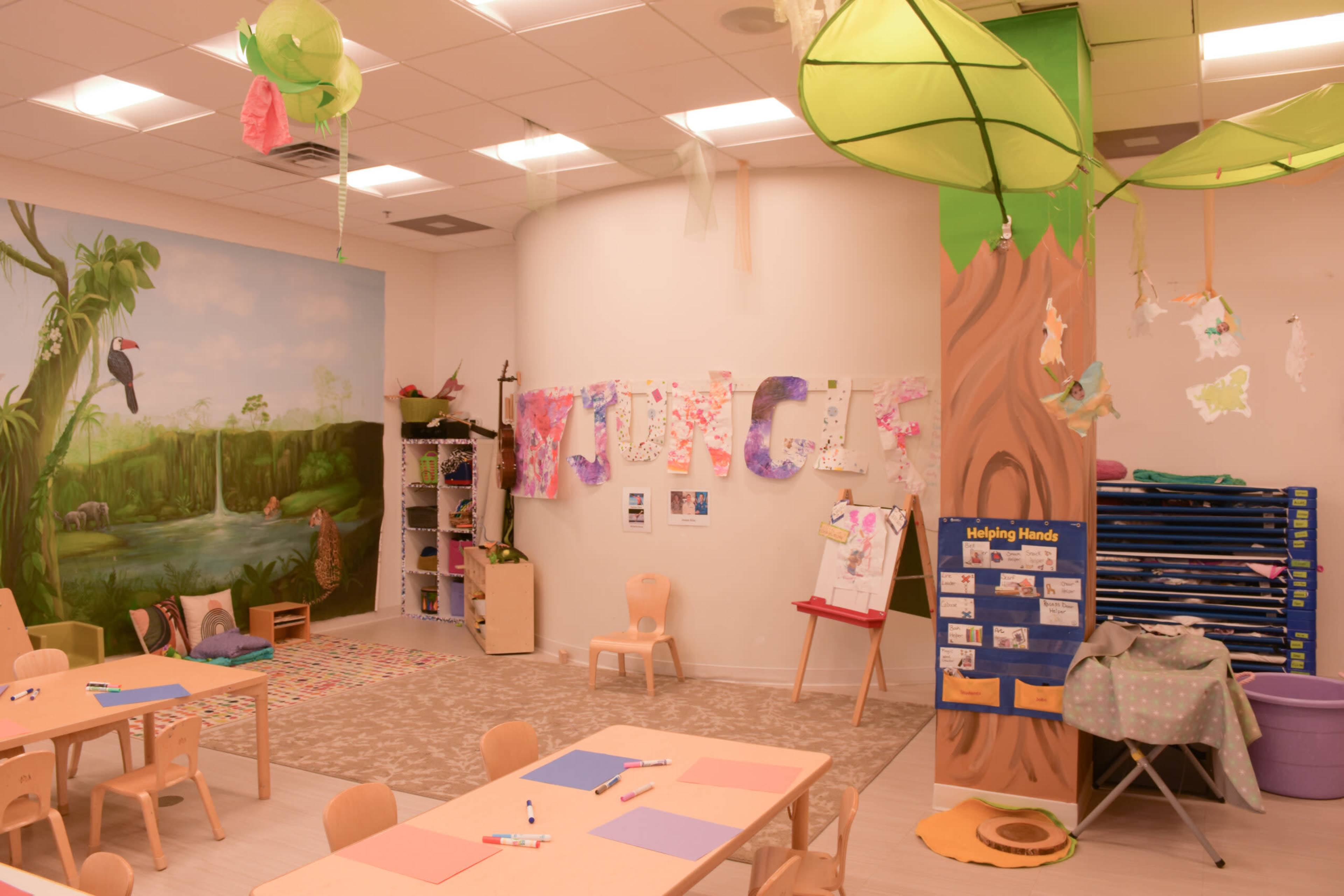 A brightly colored classroom features a mural of a waterfall and tropical plants, with tables set for activities and various art supplies and decorations hanging from the ceiling.