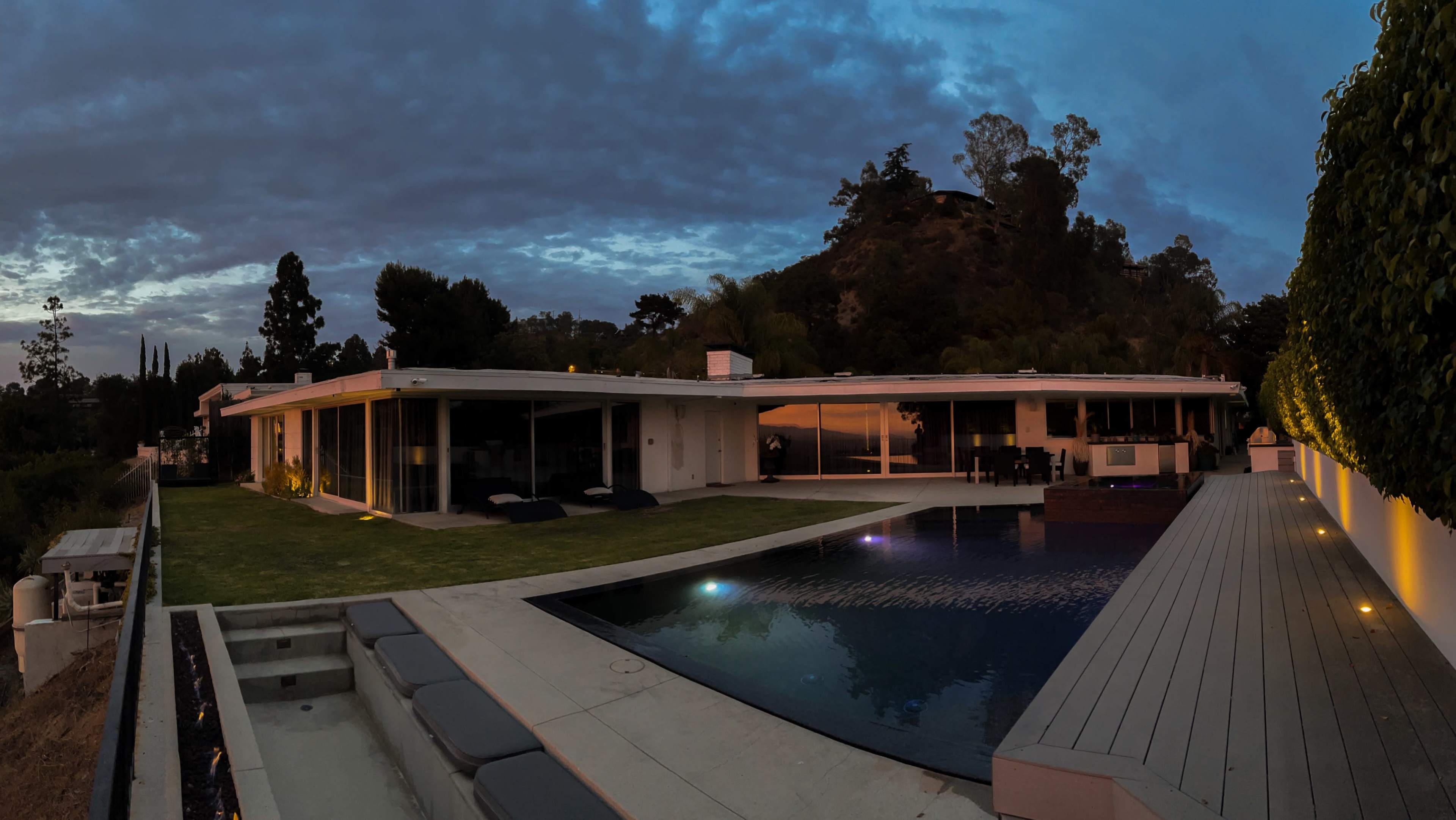 A modern house with large glass windows overlooks a swimming pool and hillside at dusk.