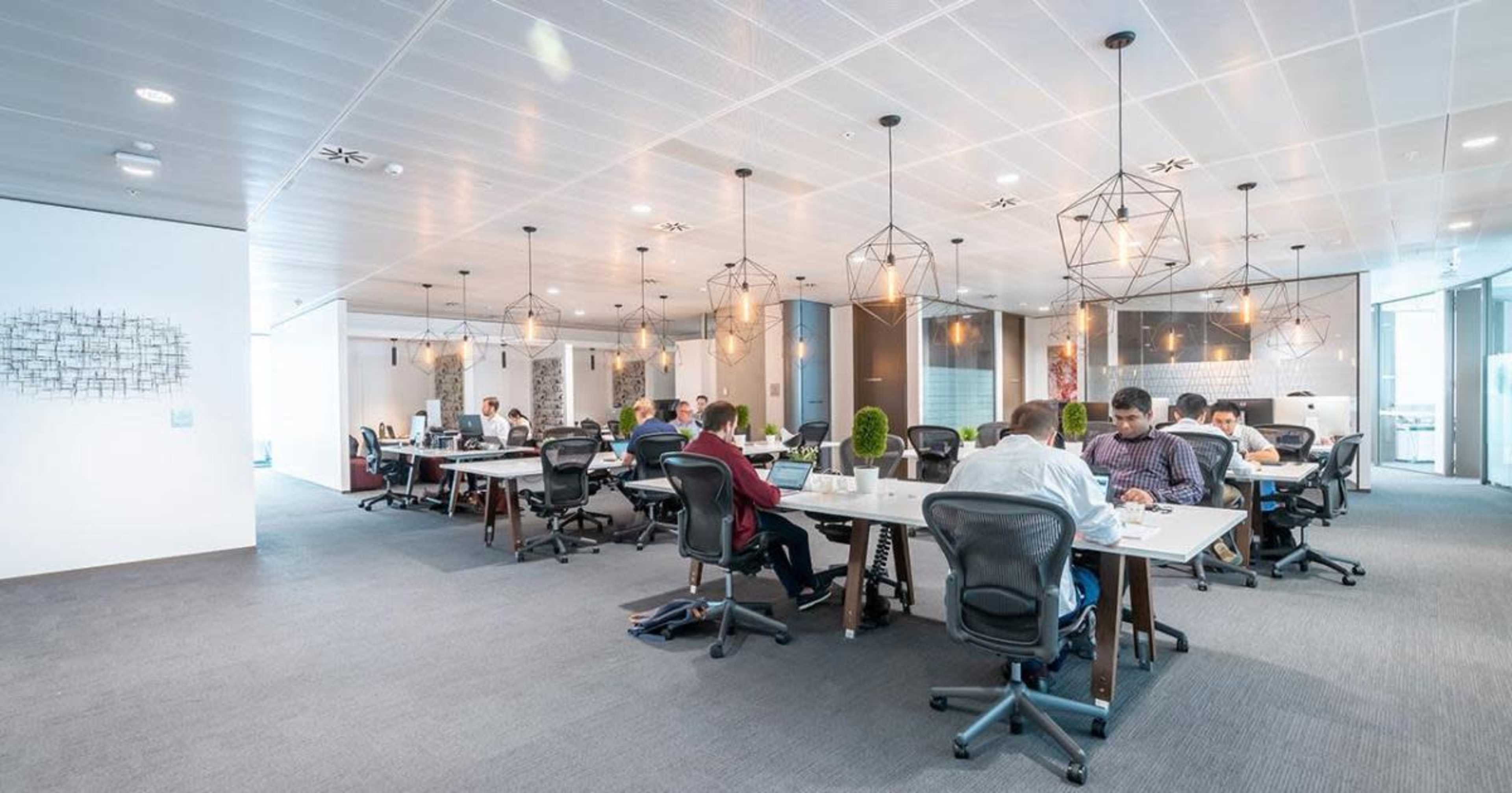 A modern office space with several workers seated at desks, engaged in various tasks, and pendant lights hanging from the ceiling.