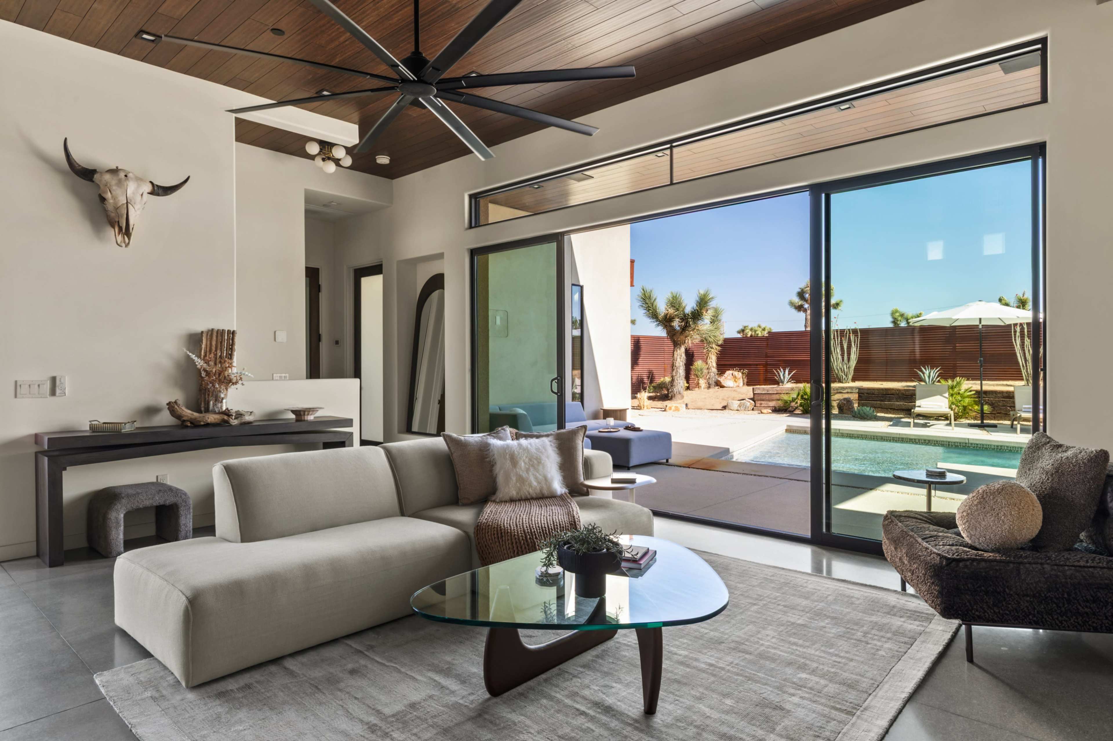 A modern living room features a spacious sectional sofa, a glass coffee table, and large windows overlooking a pool and patio area.