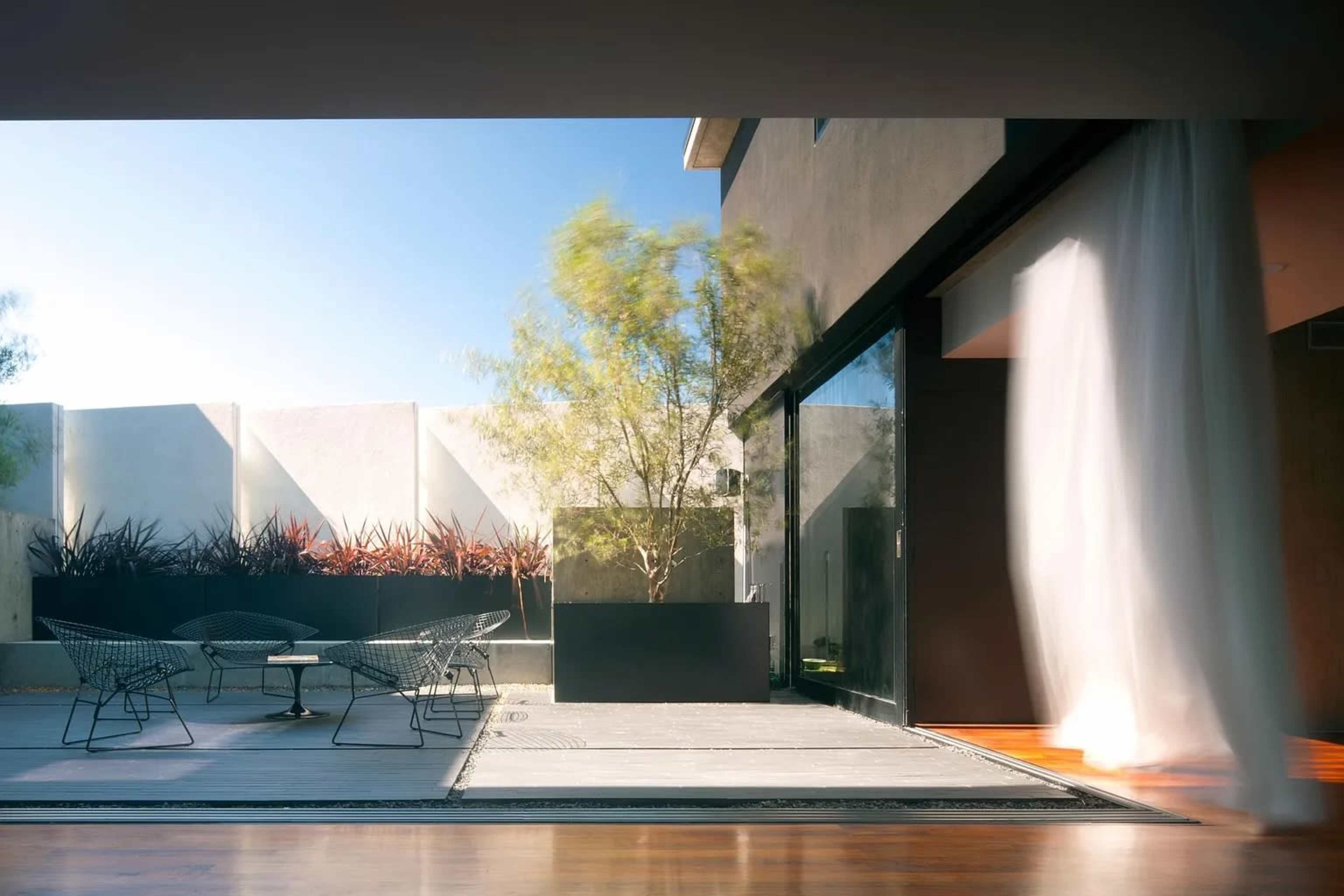 A modern patio features a small table with two chairs, bordered by plants and a textured wall under a clear blue sky.