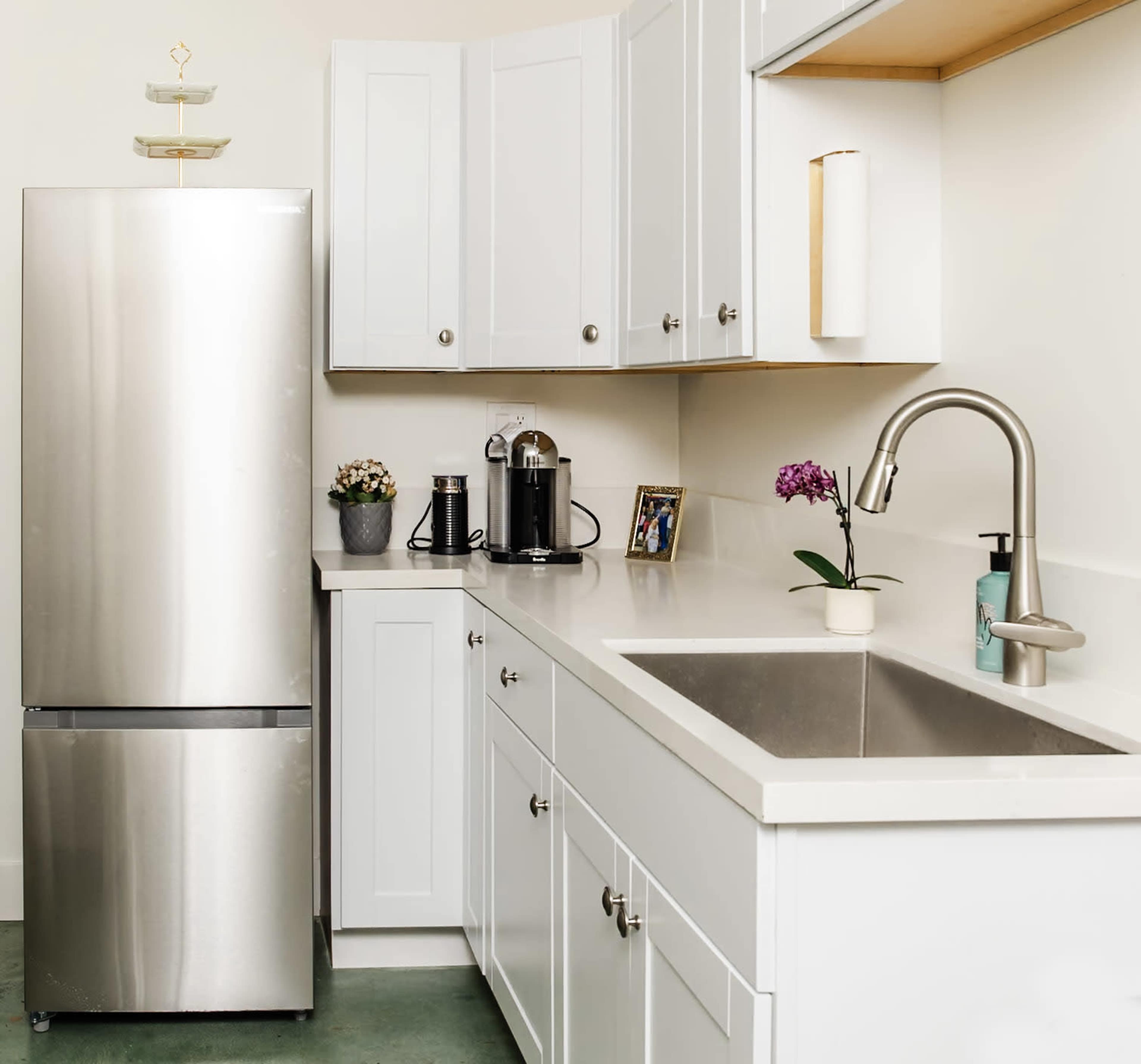 A modern kitchen features a stainless steel refrigerator, white cabinets, a countertop with a sink, and a coffee machine beside a potted plant.