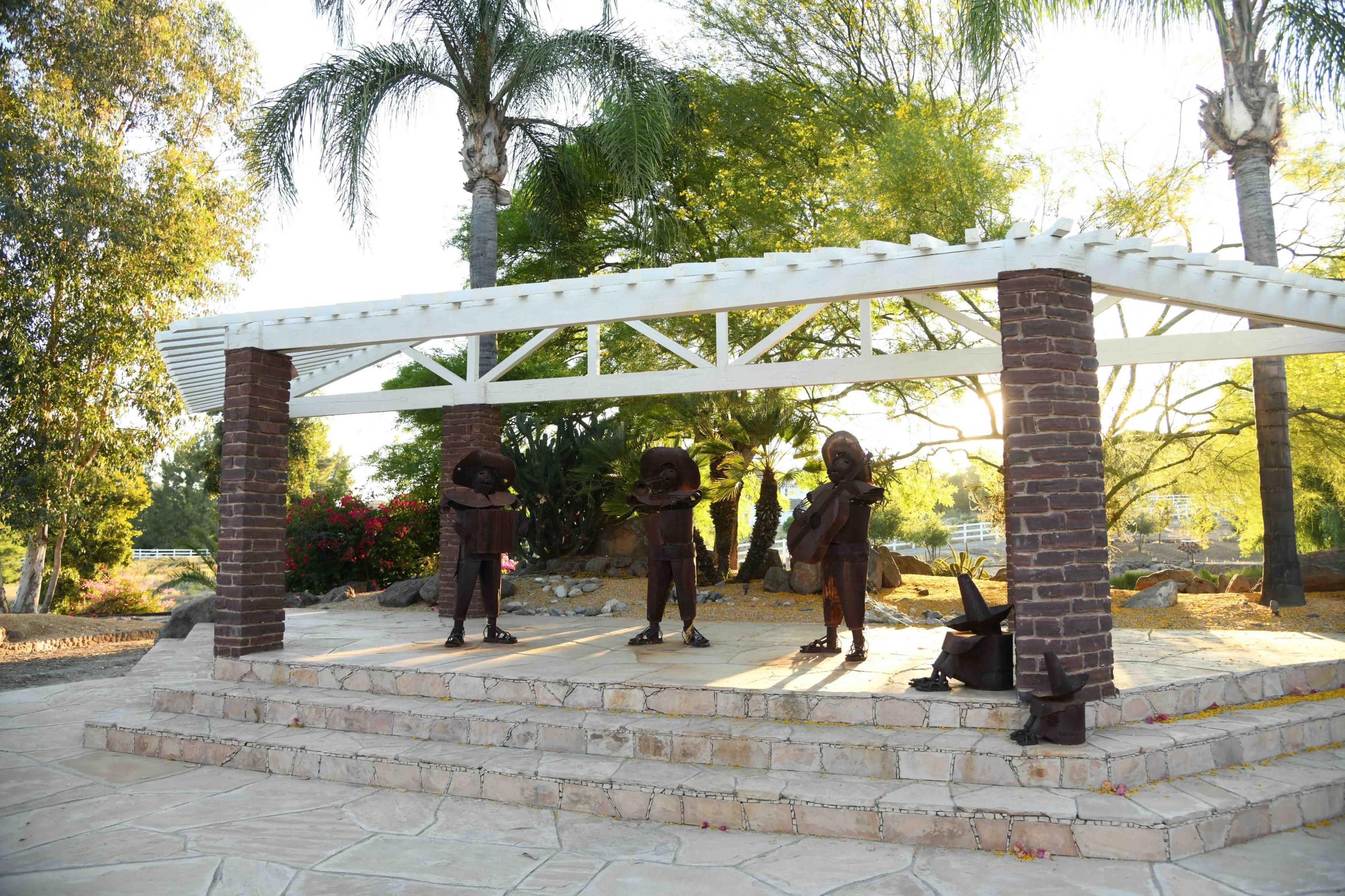 A three-figure bronze sculpture of musicians stands under a white gazebo surrounded by trees and landscaping.