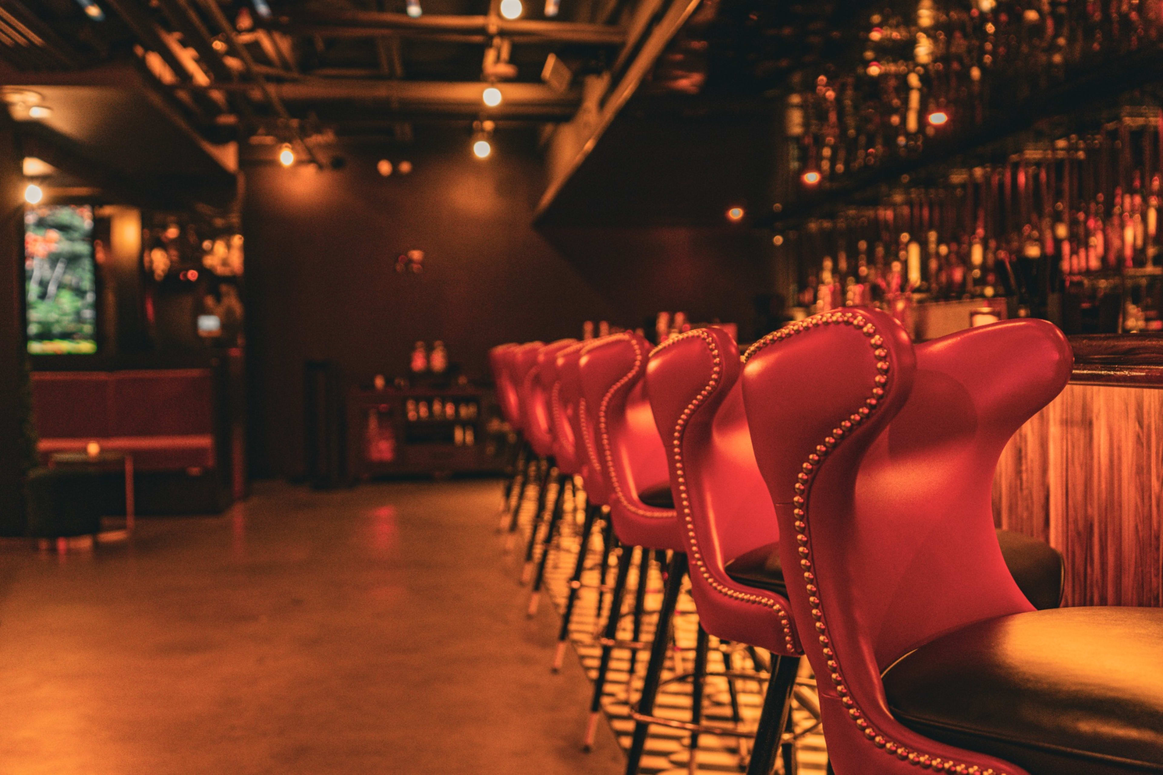 The image shows an interior bar scene featuring a row of red upholstered bar stools in front of a wooden bar counter.