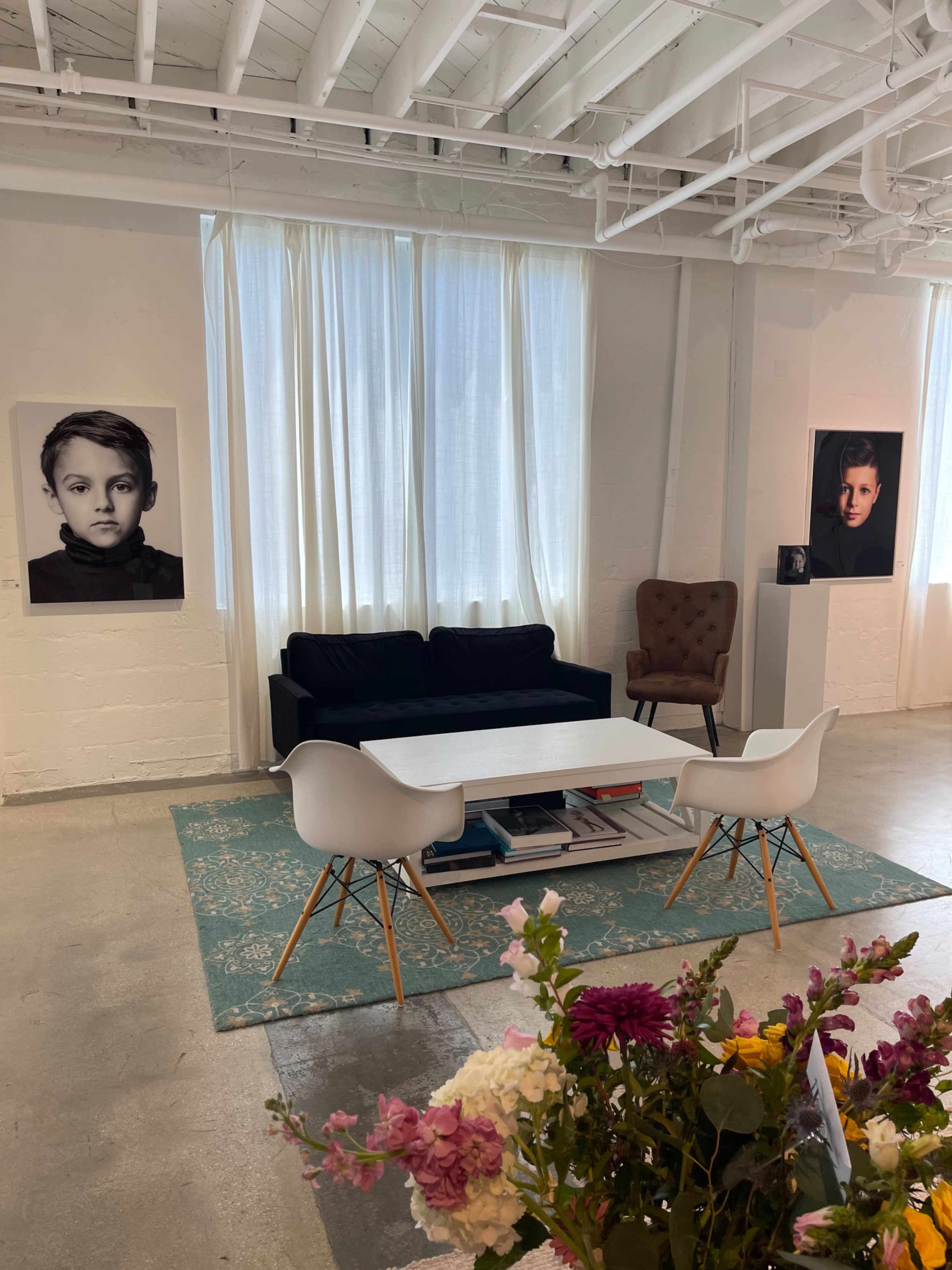 The image shows a bright gallery-like space with a black sofa, a white table, and large portraits of children on the walls, complemented by a floral arrangement in the foreground.