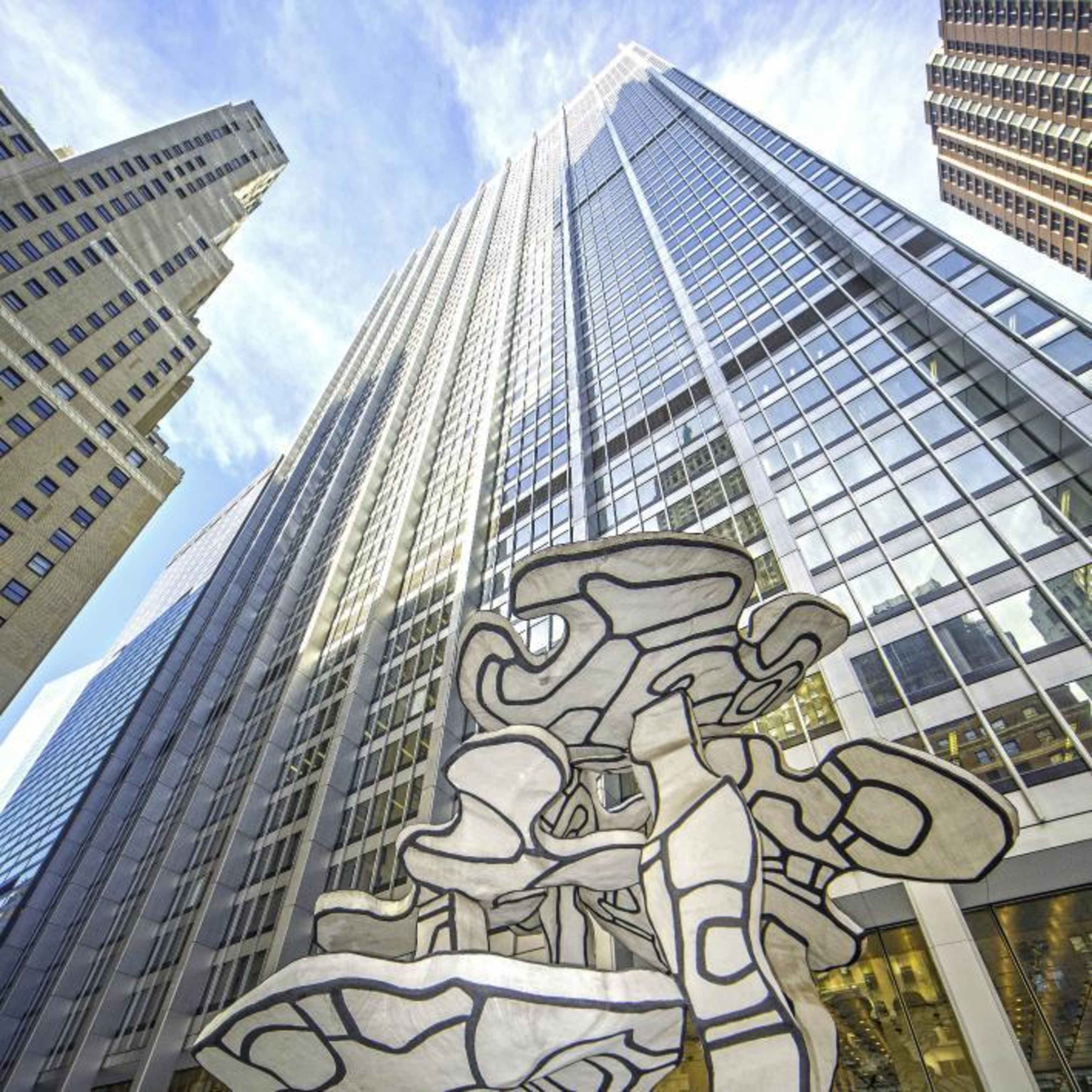 A large abstract sculpture stands in the foreground of a towering glass skyscraper, with a clear blue sky above.