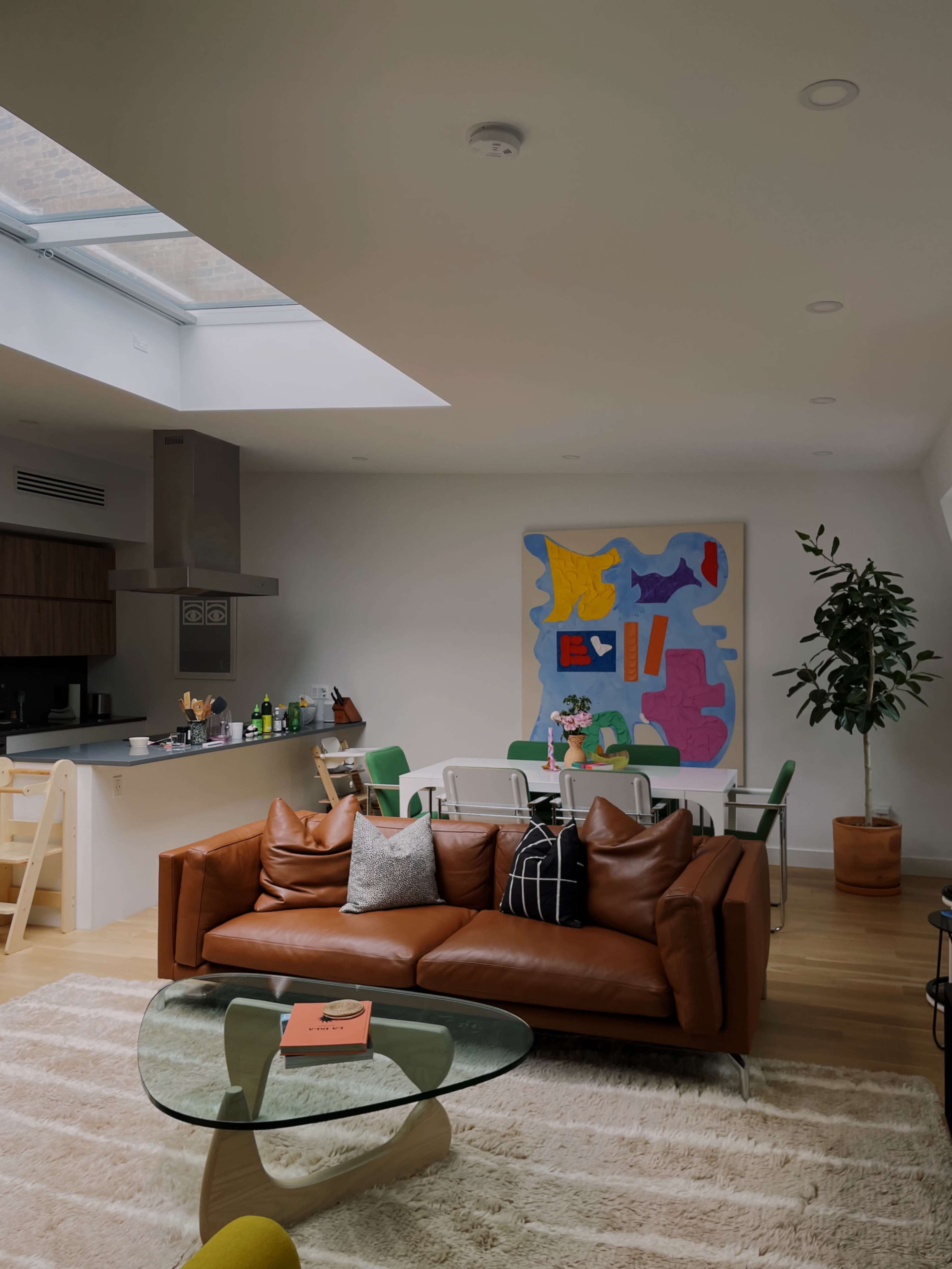 The image shows a modern living space featuring a brown leather sofa, a glass coffee table, and a dining area with green chairs, illuminated by a skylight.
