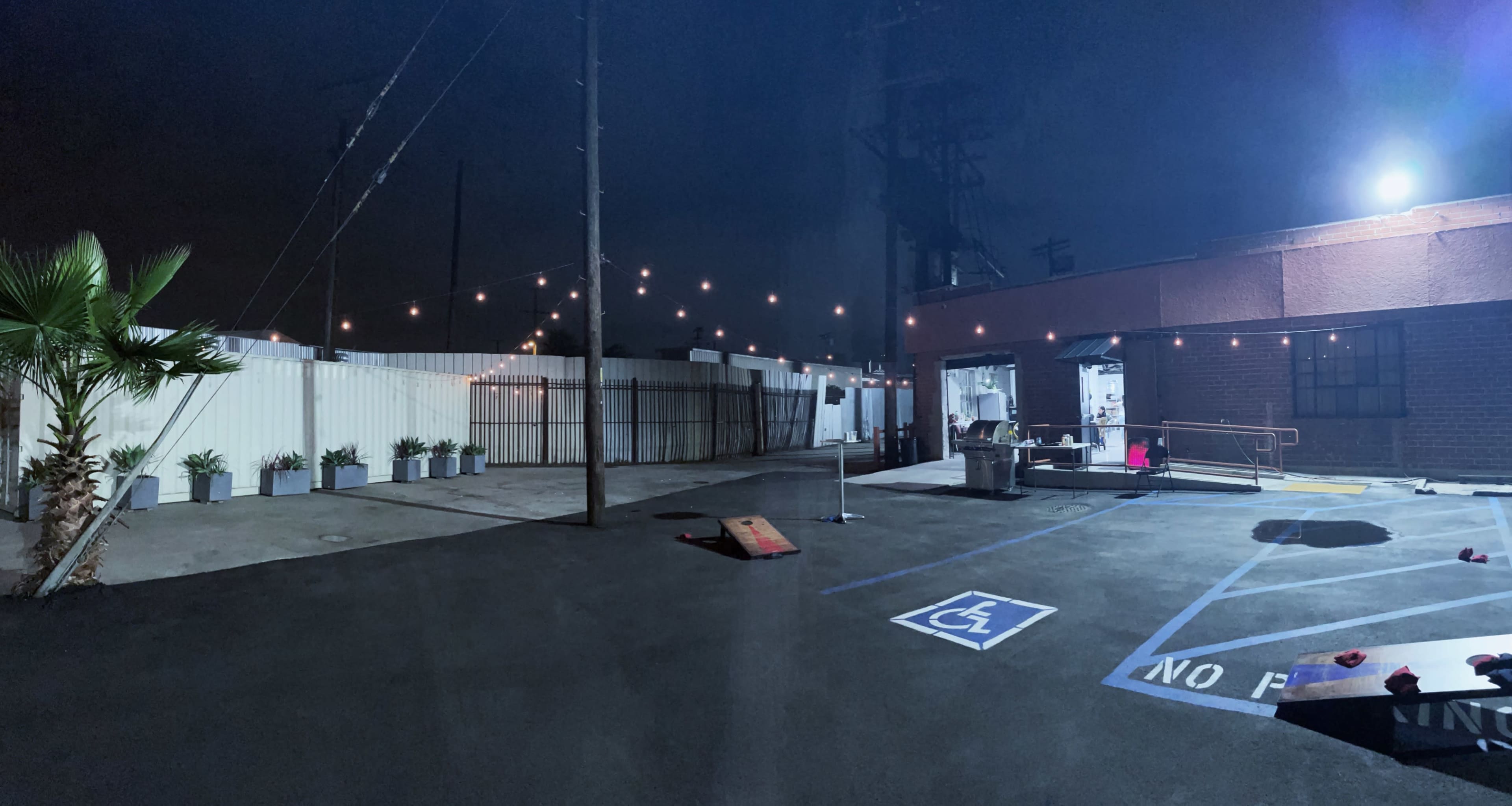A dimly lit outdoor area adjacent to a building, featuring string lights overhead, a marked wheelchair-accessible parking space, and a few potted plants lining the perimeter.