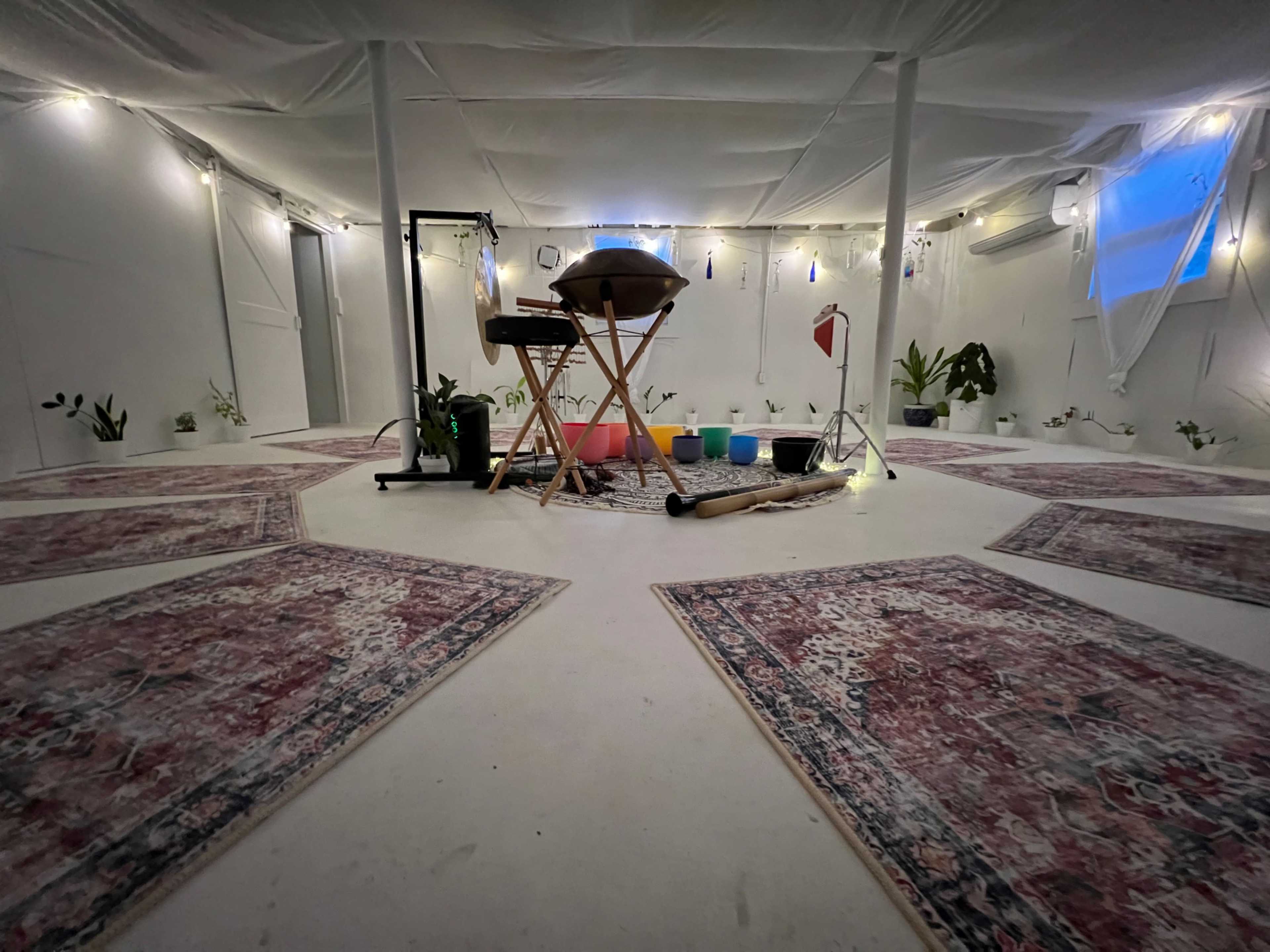 A spacious white tent interior with patterned rugs on the floor, surrounded by plants, and featuring a central setup with a wooden table, colorful cushions, and ambient lighting.