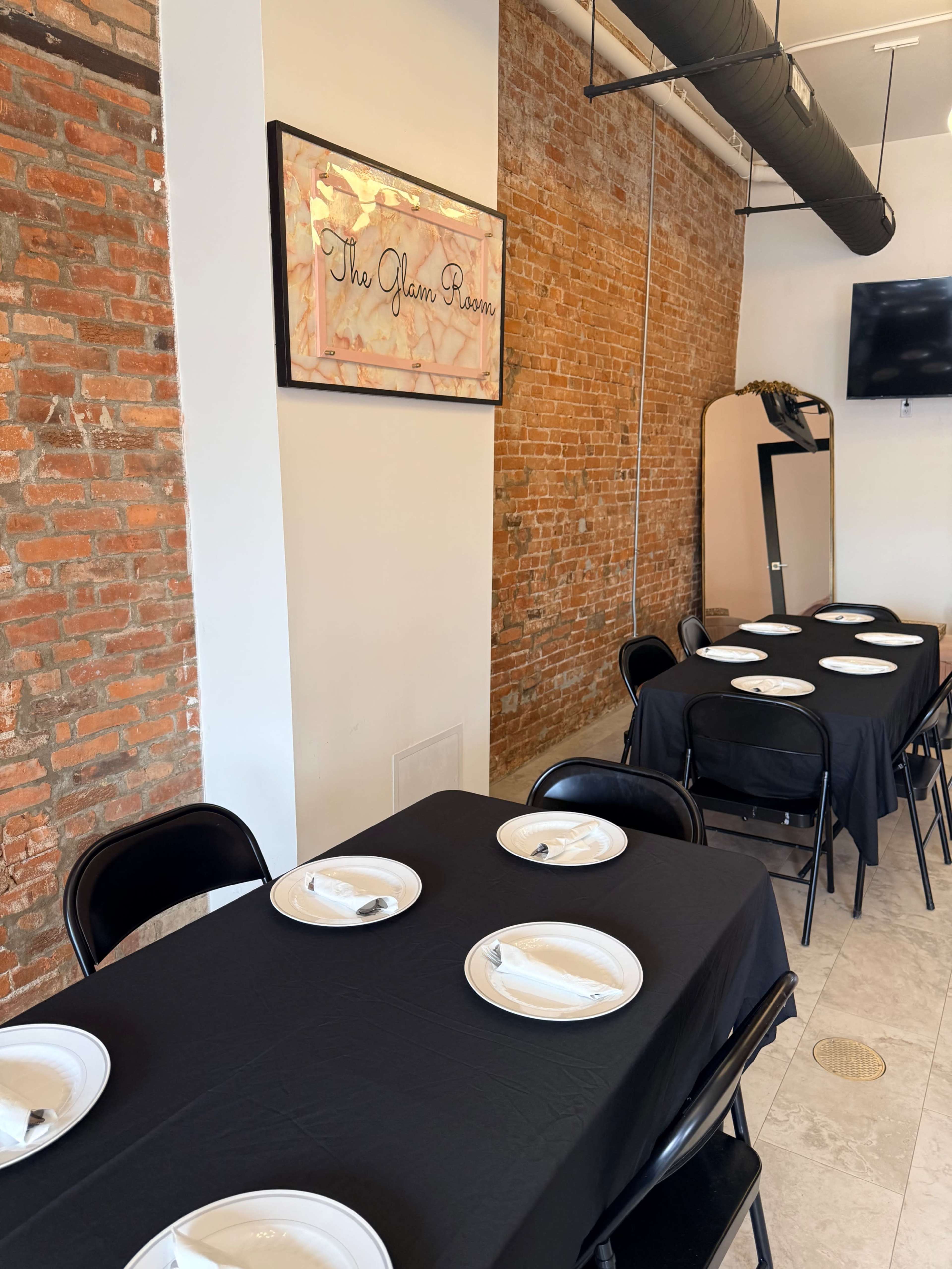 The image shows a dining area with several black-covered tables, white plates set at each place, and a brick wall adorned with a sign that reads "The Glam Room."