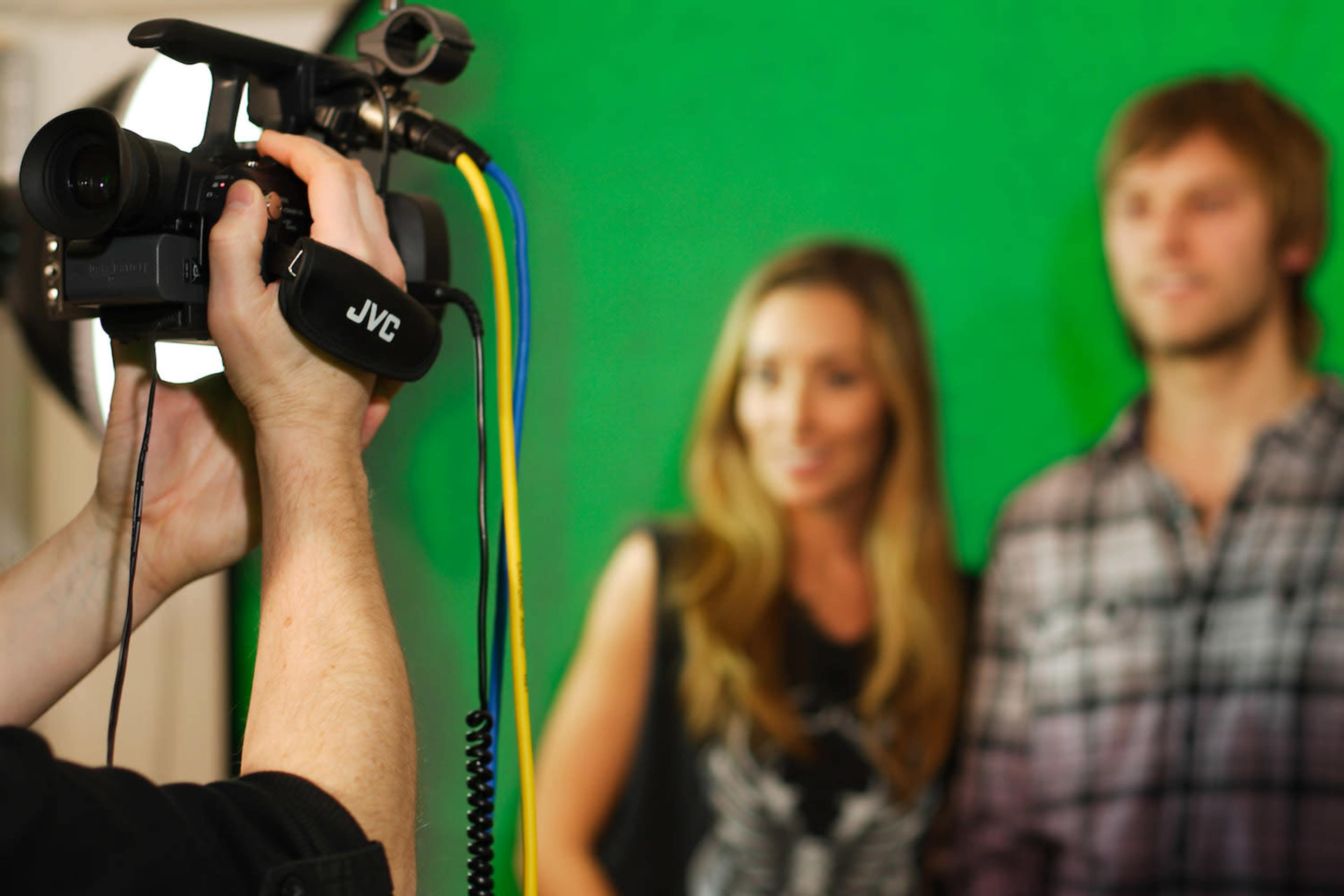 A person is holding a camera while recording two individuals in front of a green screen.