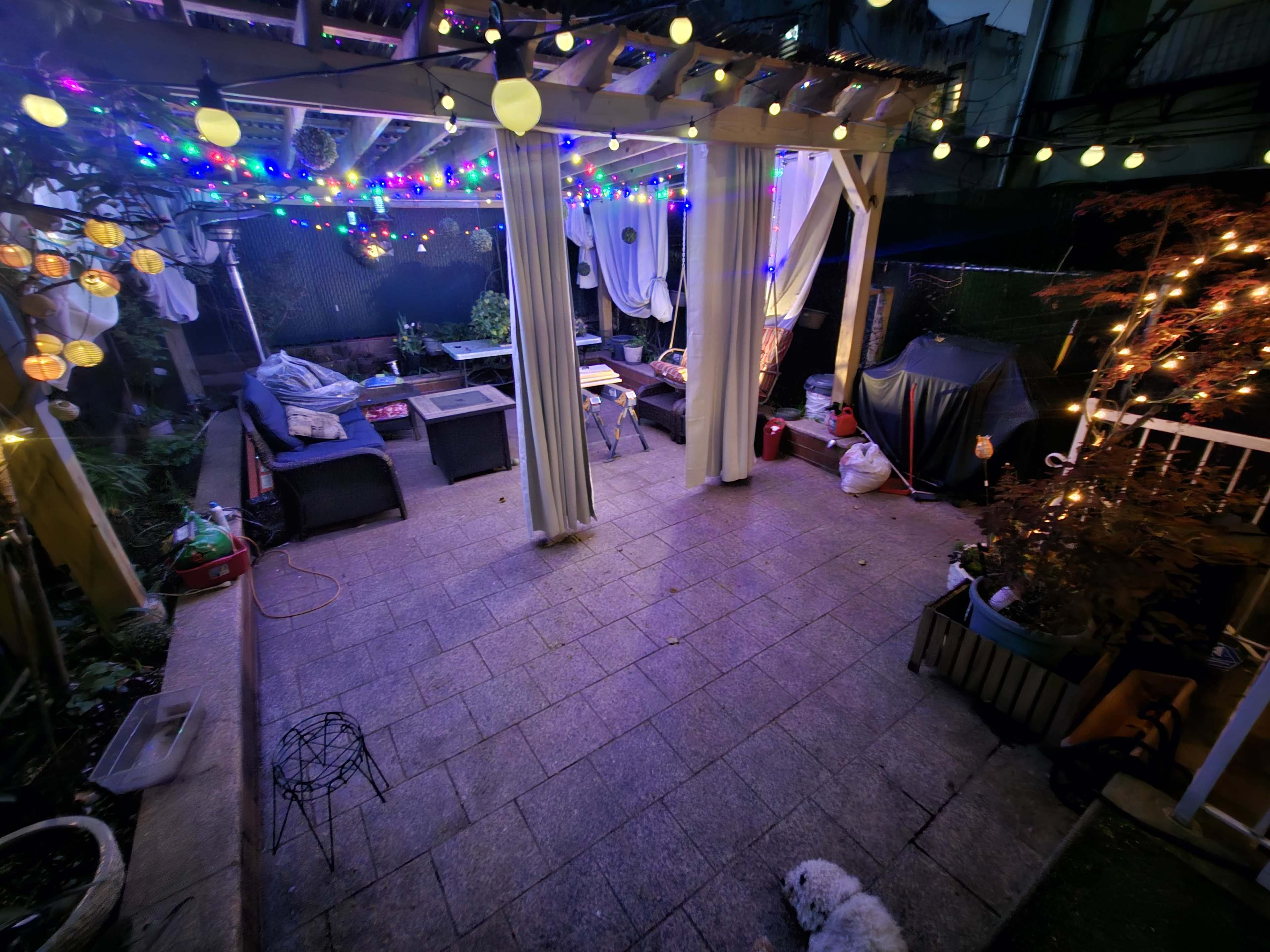 A patio is decorated with colorful string lights and features outdoor furniture, while a small dog is lying on the ground.
