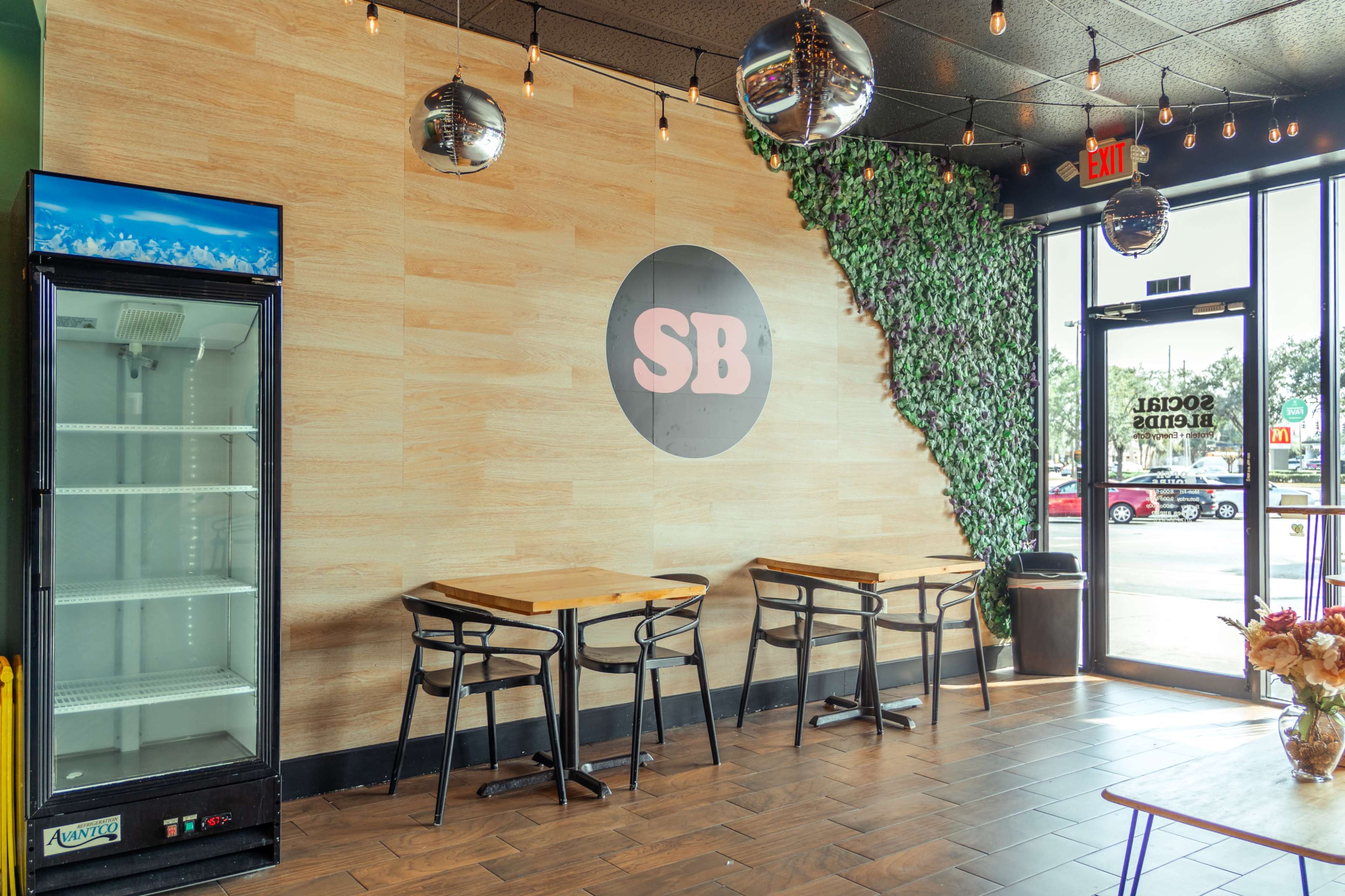 The image shows a cafe interior featuring wooden tables and chairs, a wall adorned with greenery, and a large circular logo on the wall.