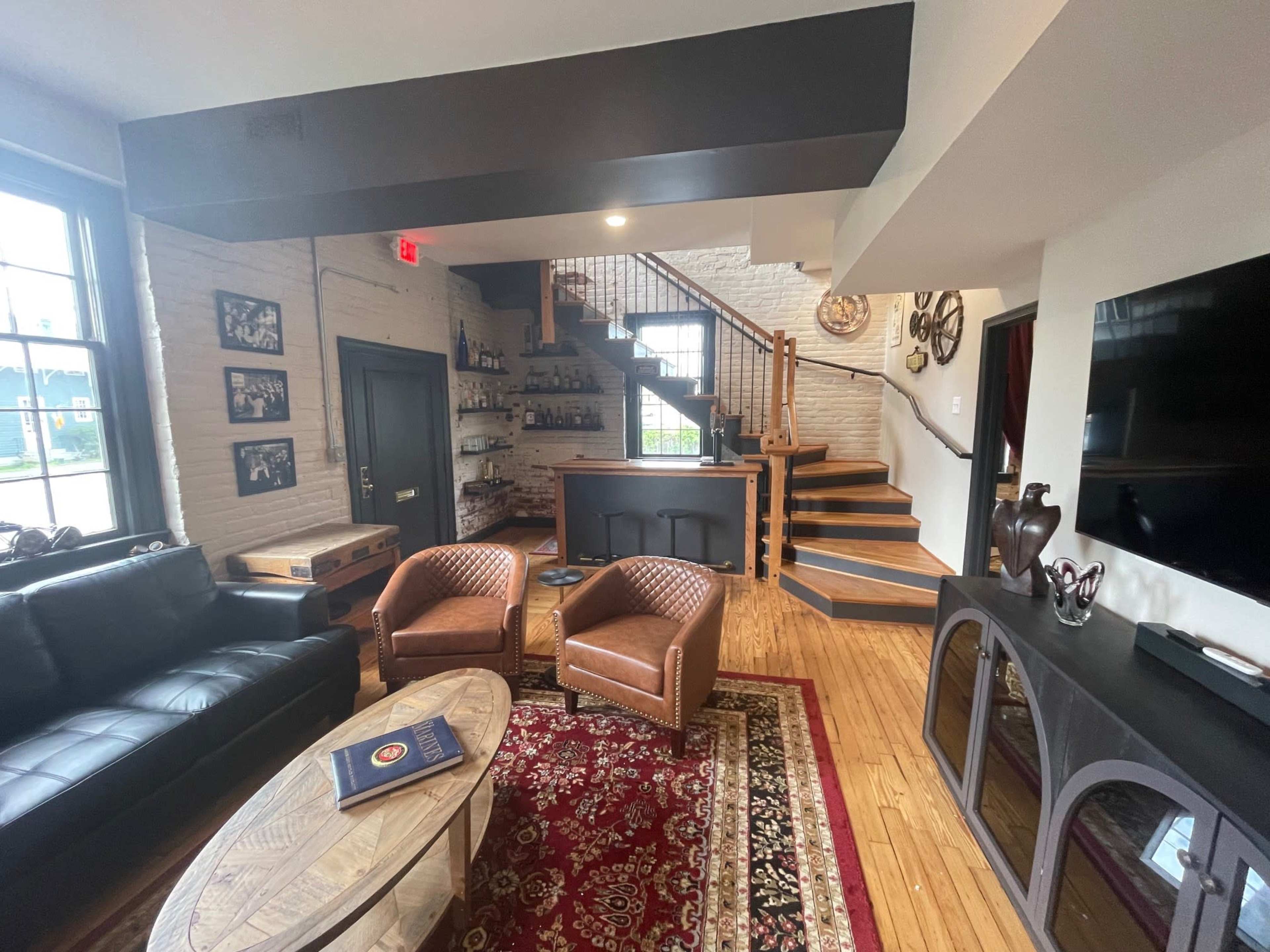 The image shows a modern living area featuring a black leather sofa, two brown armchairs, a round coffee table on a patterned rug, and a staircase leading to another level.