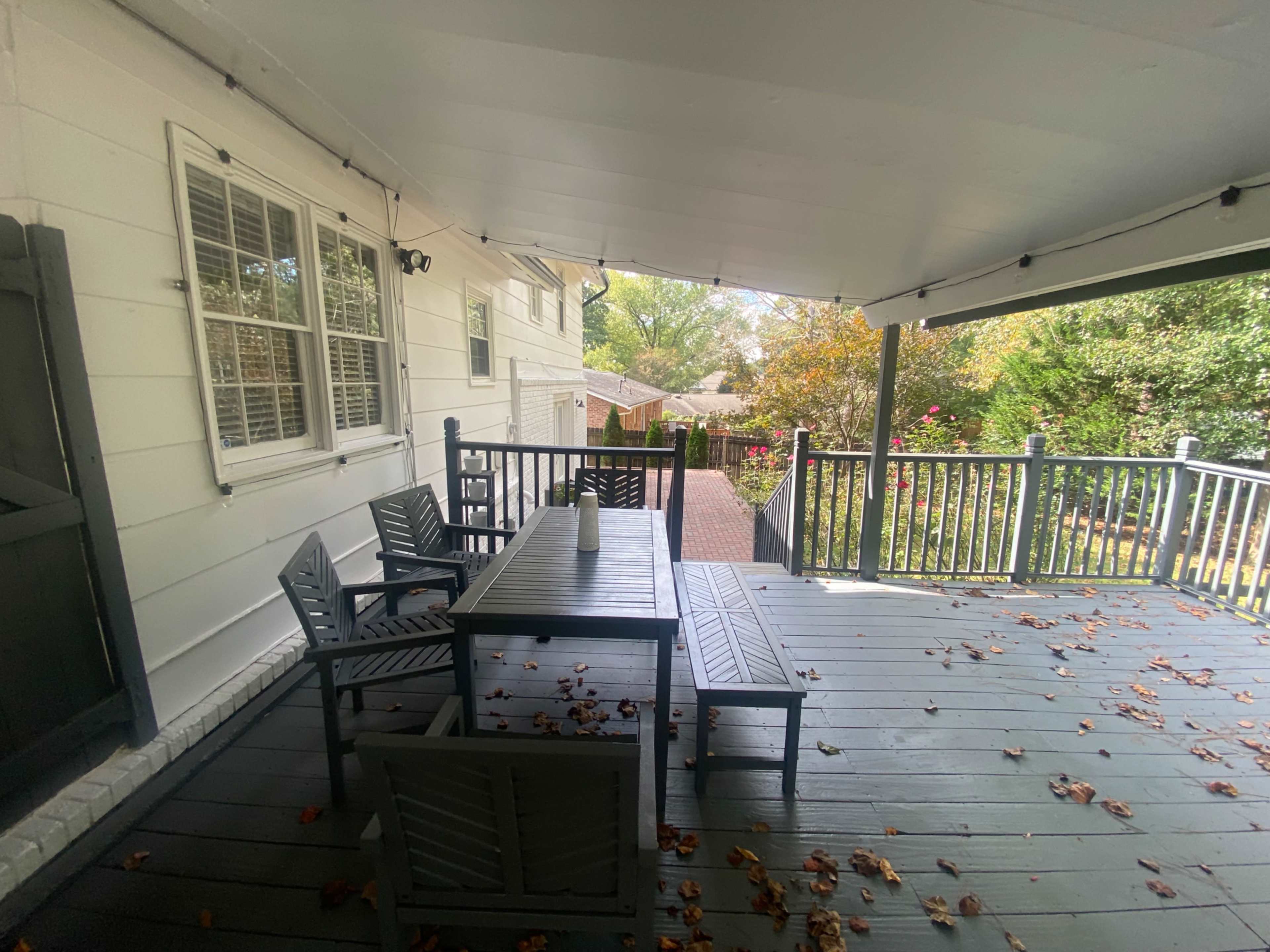 A covered deck features a wooden table and chairs surrounded by fallen leaves, with a view of a yard and garden beyond.