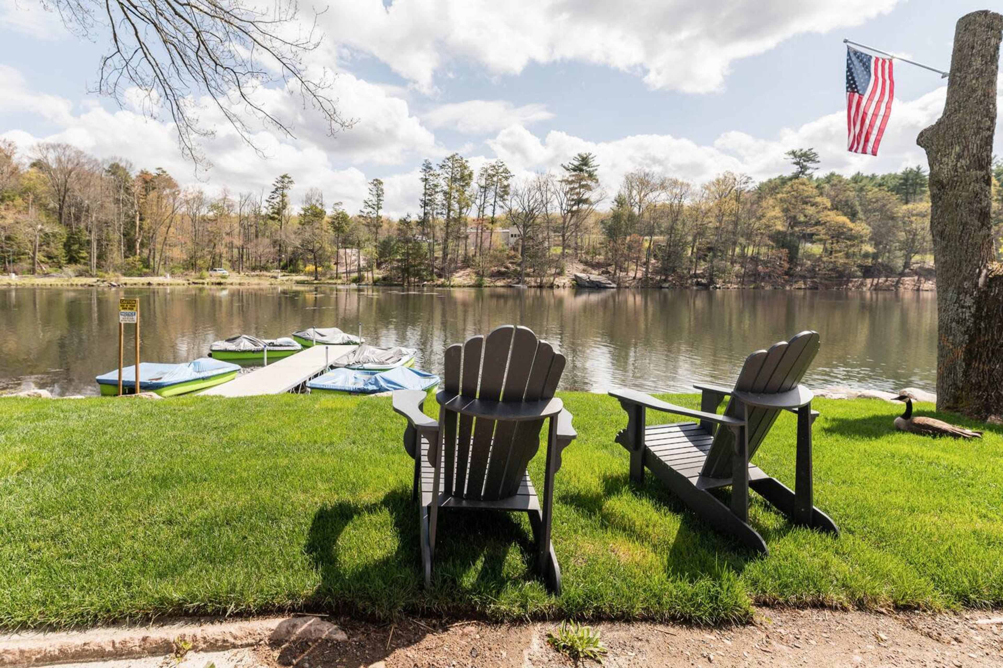 Two black adirondack chairs sit on green grass beside a calm lake, with a boat dock and an American flag in the background.