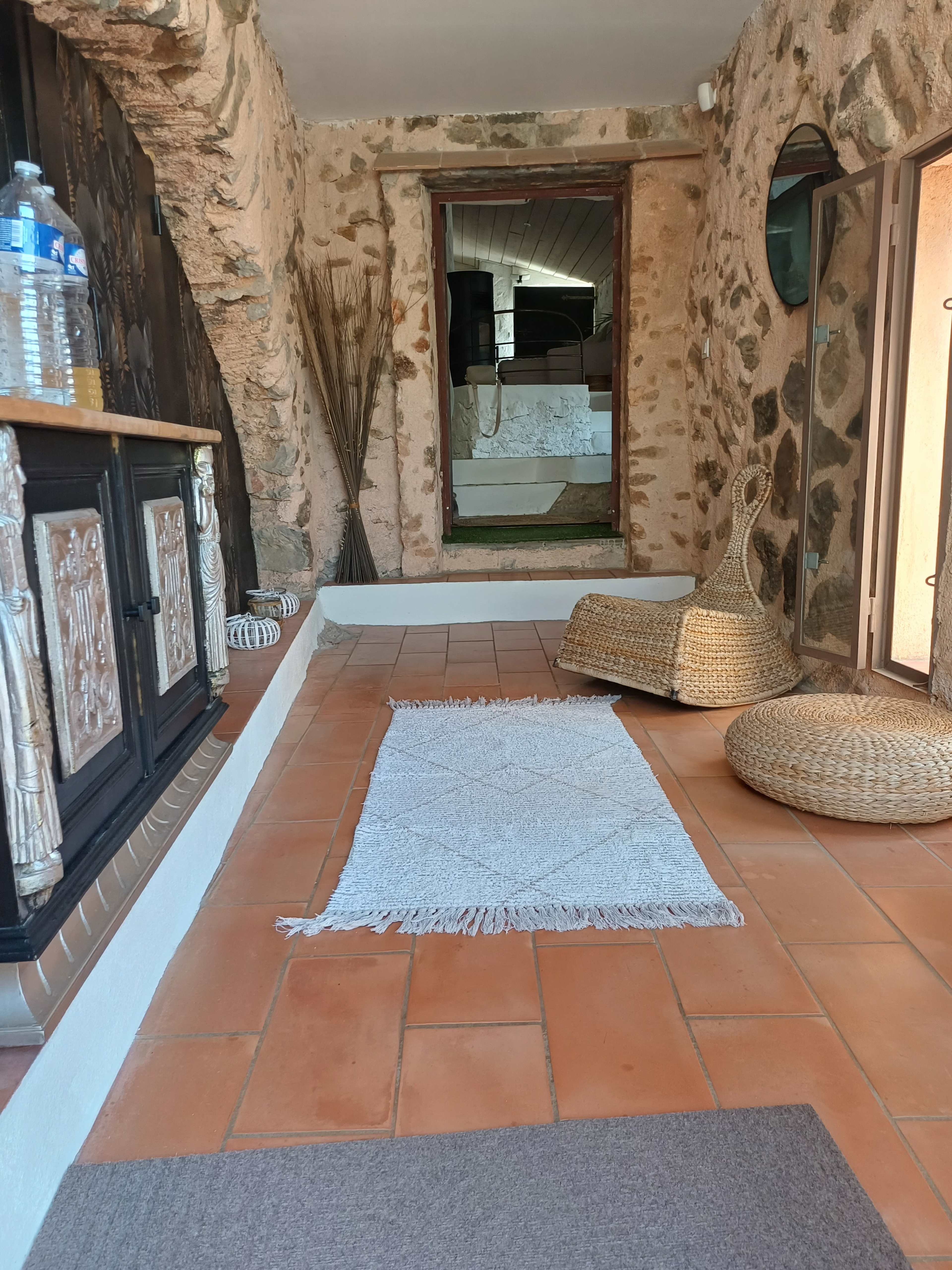 The image shows a cozy entryway with terracotta tiles, stone walls, a woven rug, and decorative elements like a wooden cabinet and wicker furniture.