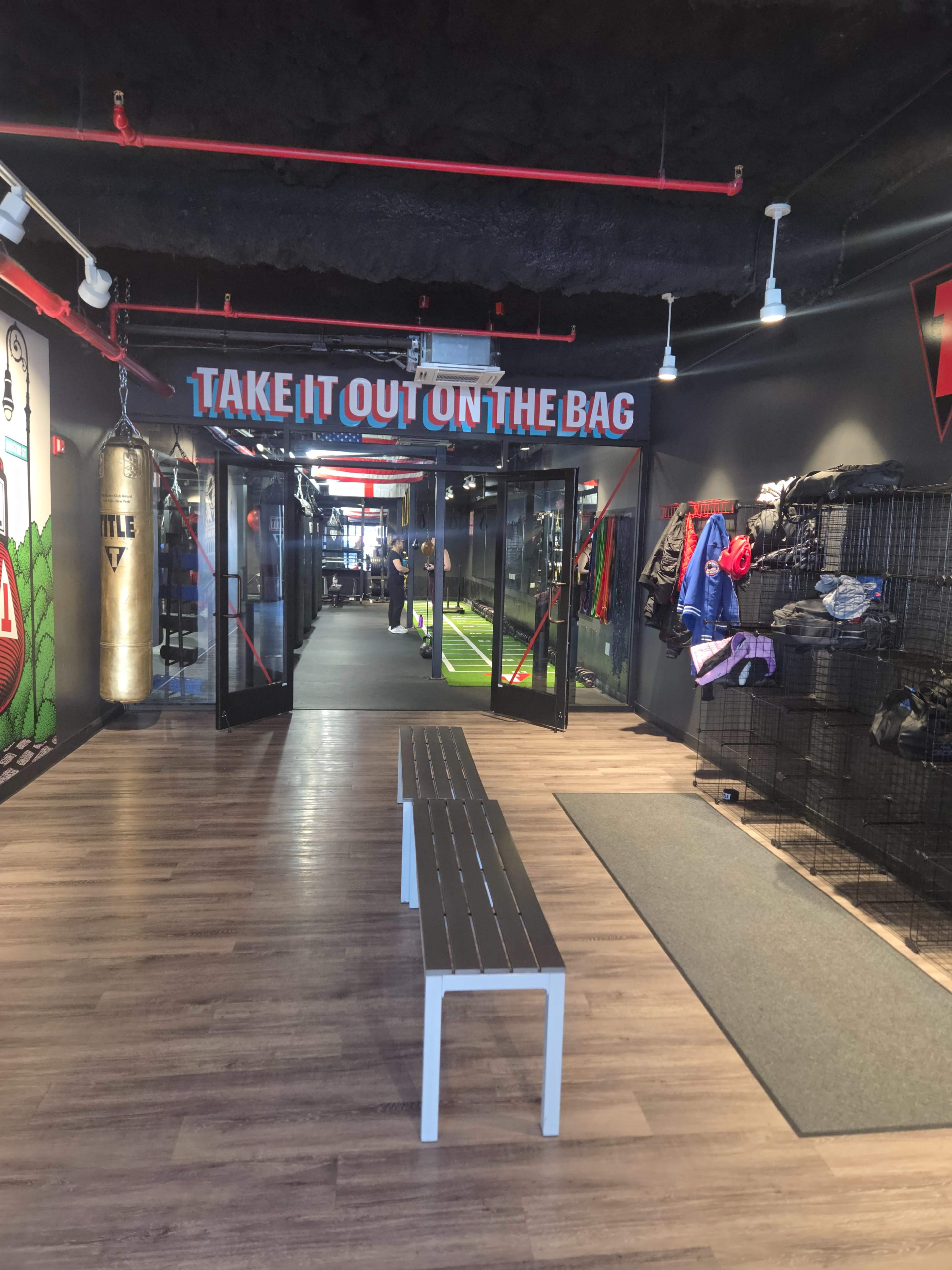 The image shows the entrance of a fitness facility featuring a sign that reads "TAKE IT OUT ON THE BAG," with boxing equipment and workout gear displayed on the walls.