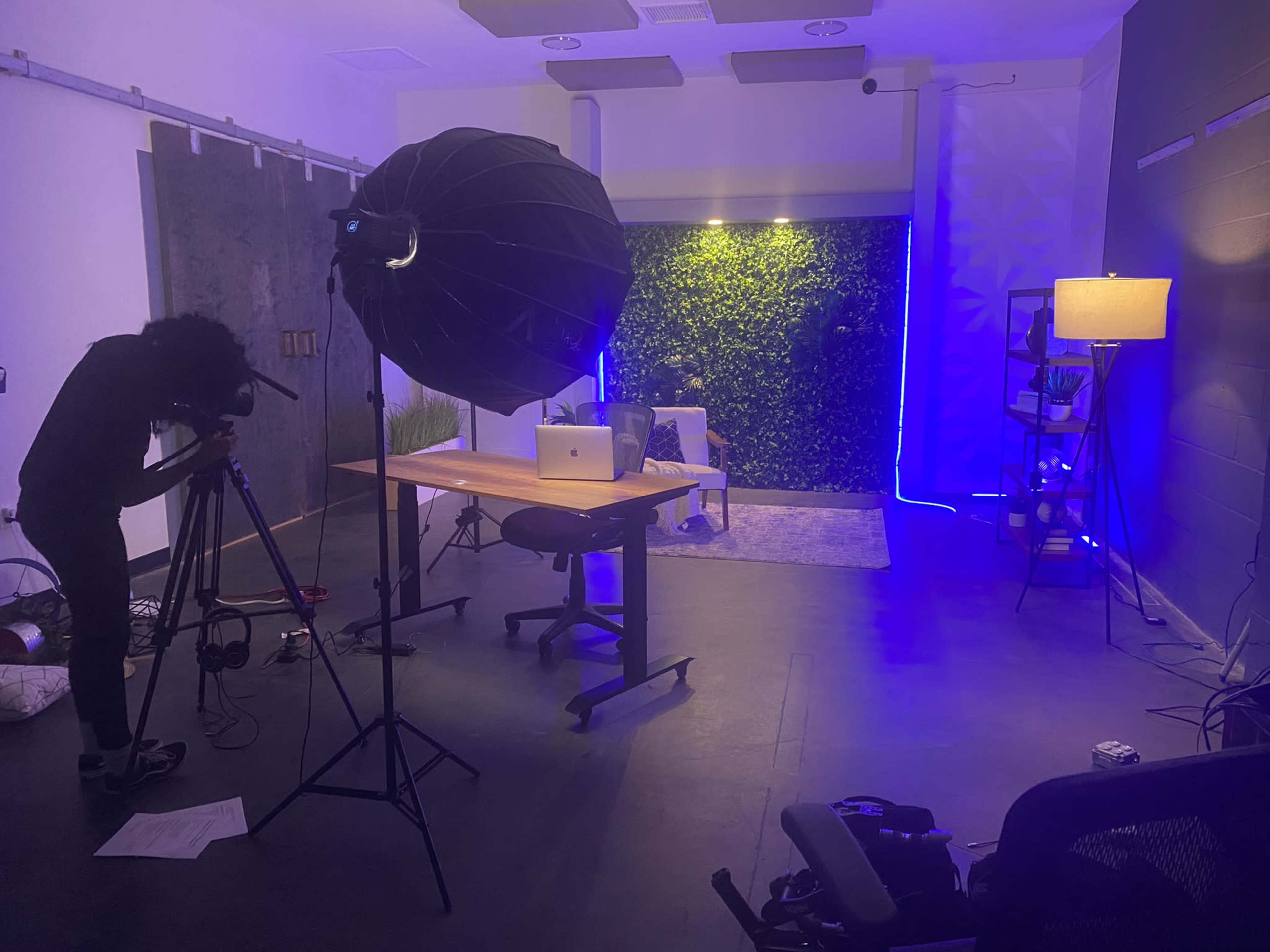 A photographer sets up a camera and lighting in a modern studio with a desk, laptop, and a greenery backdrop.