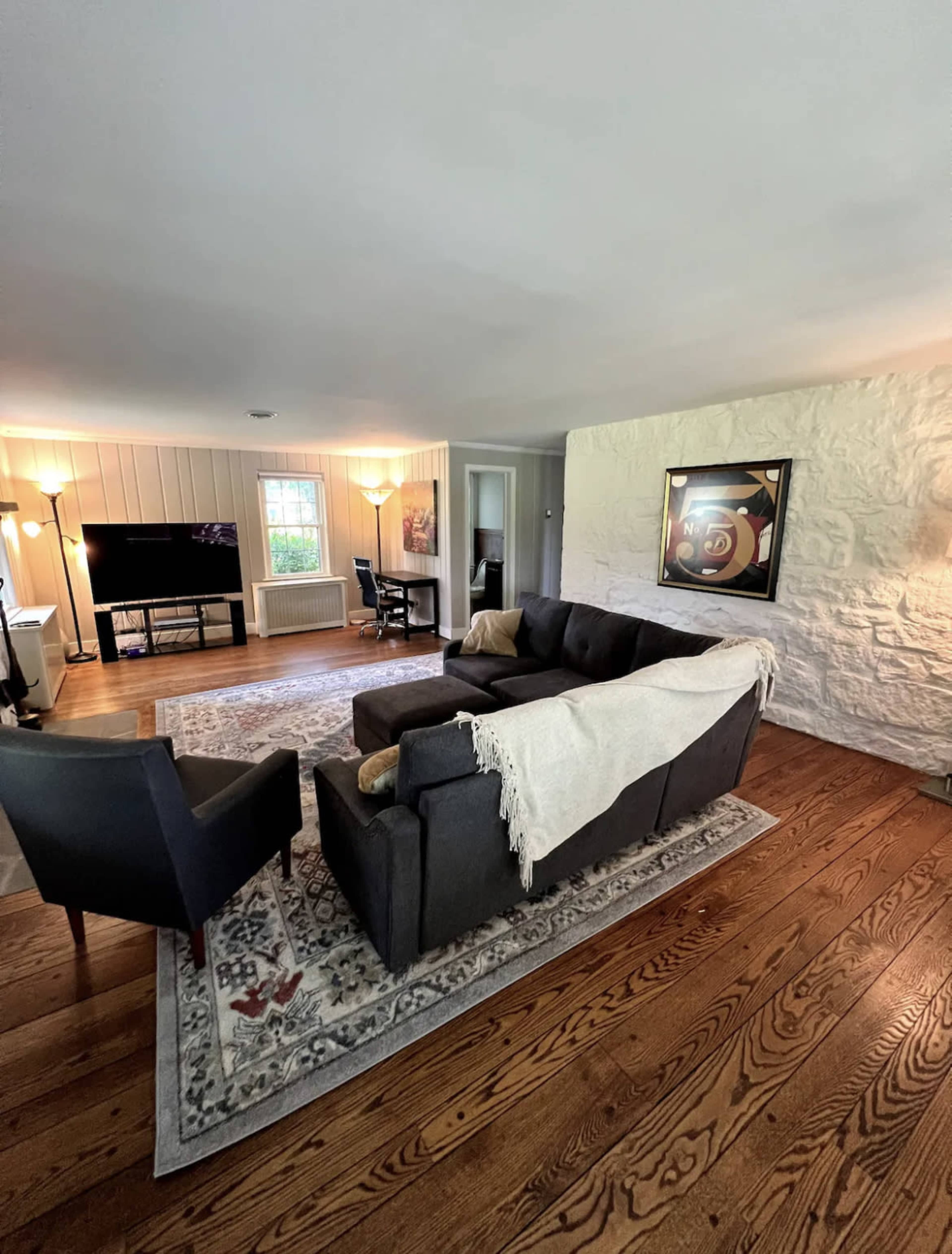 A living room with a dark sofa and armchair positioned on a wooden floor, a large area rug, a television, and a stone wall.