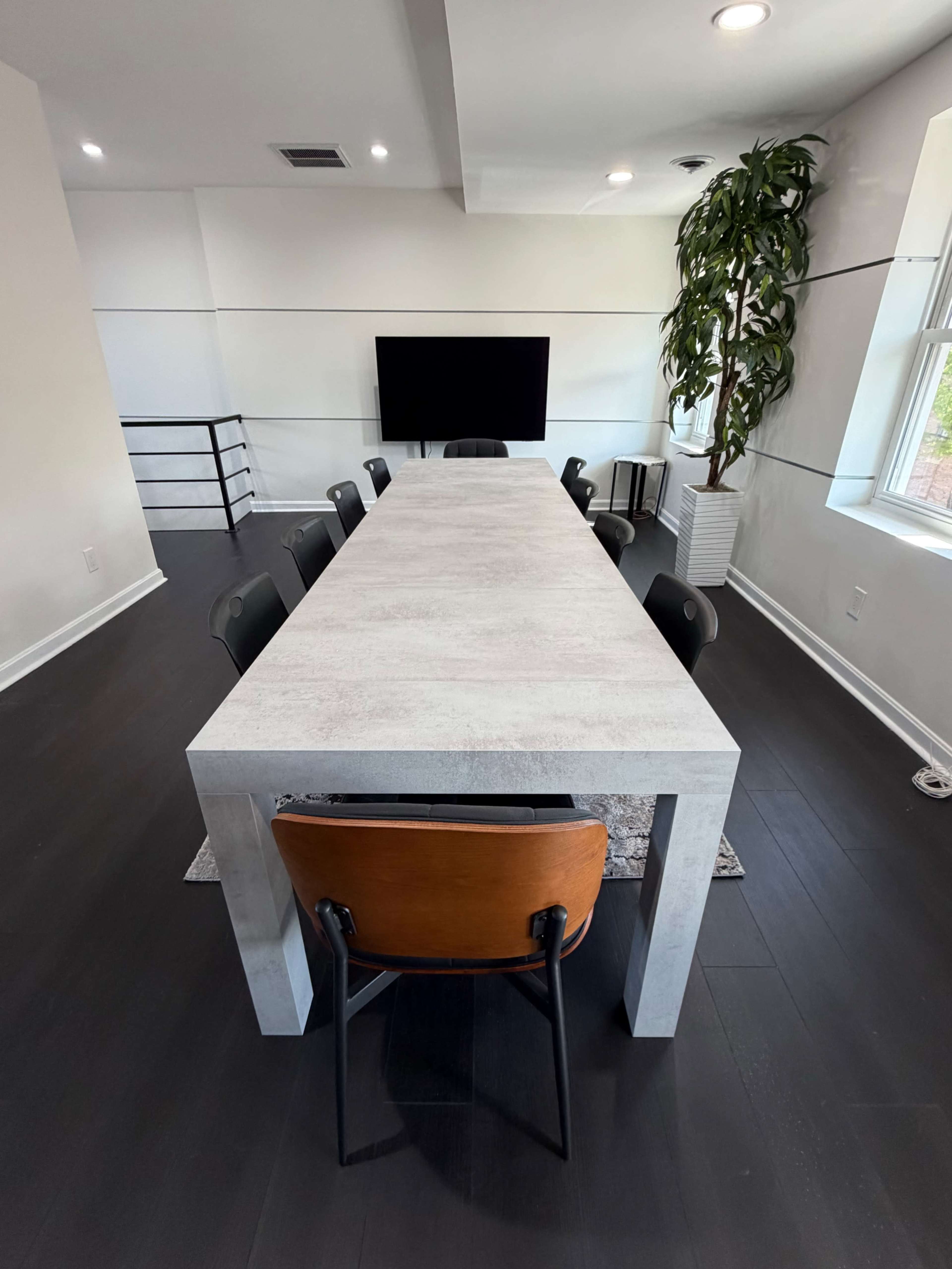 A long, modern dining table with black and wooden chairs is set in a bright, minimalistic room featuring a large TV and a decorative plant.