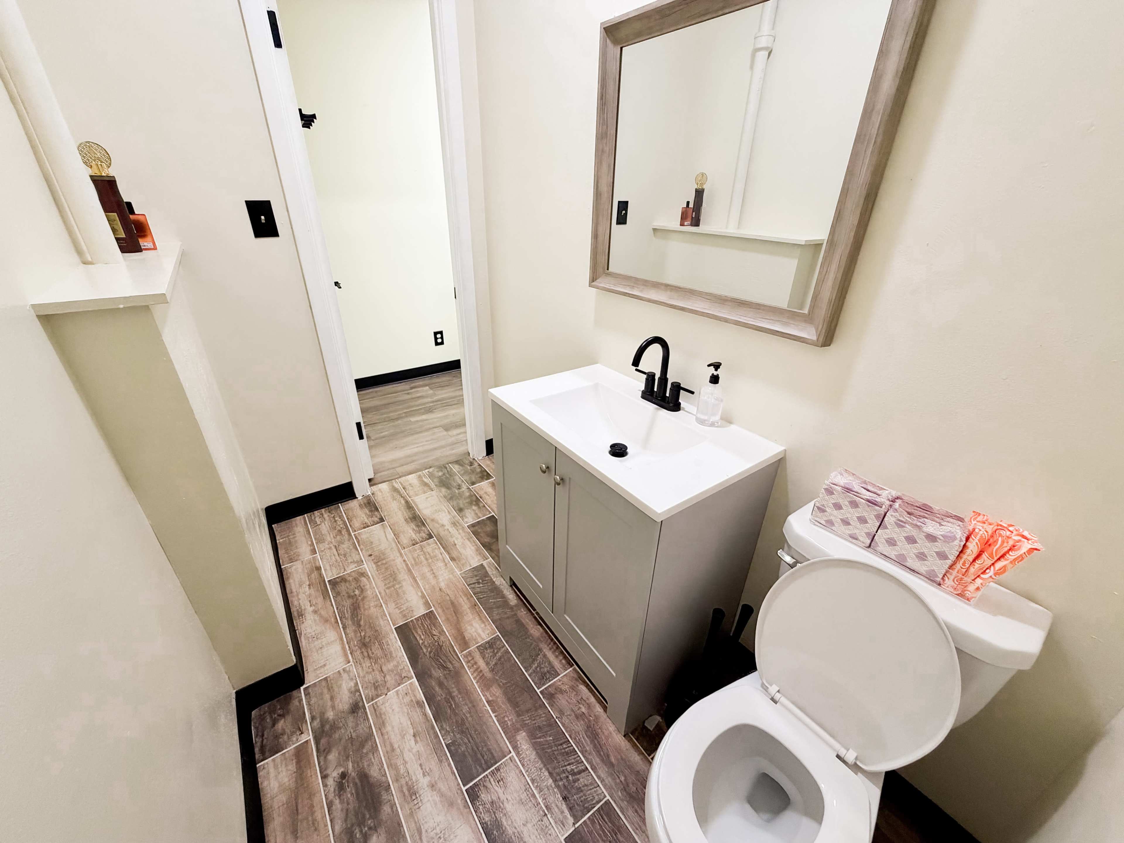 The image shows a small bathroom featuring a white sink cabinet with a mirror above it, a toilet, and tiled flooring.