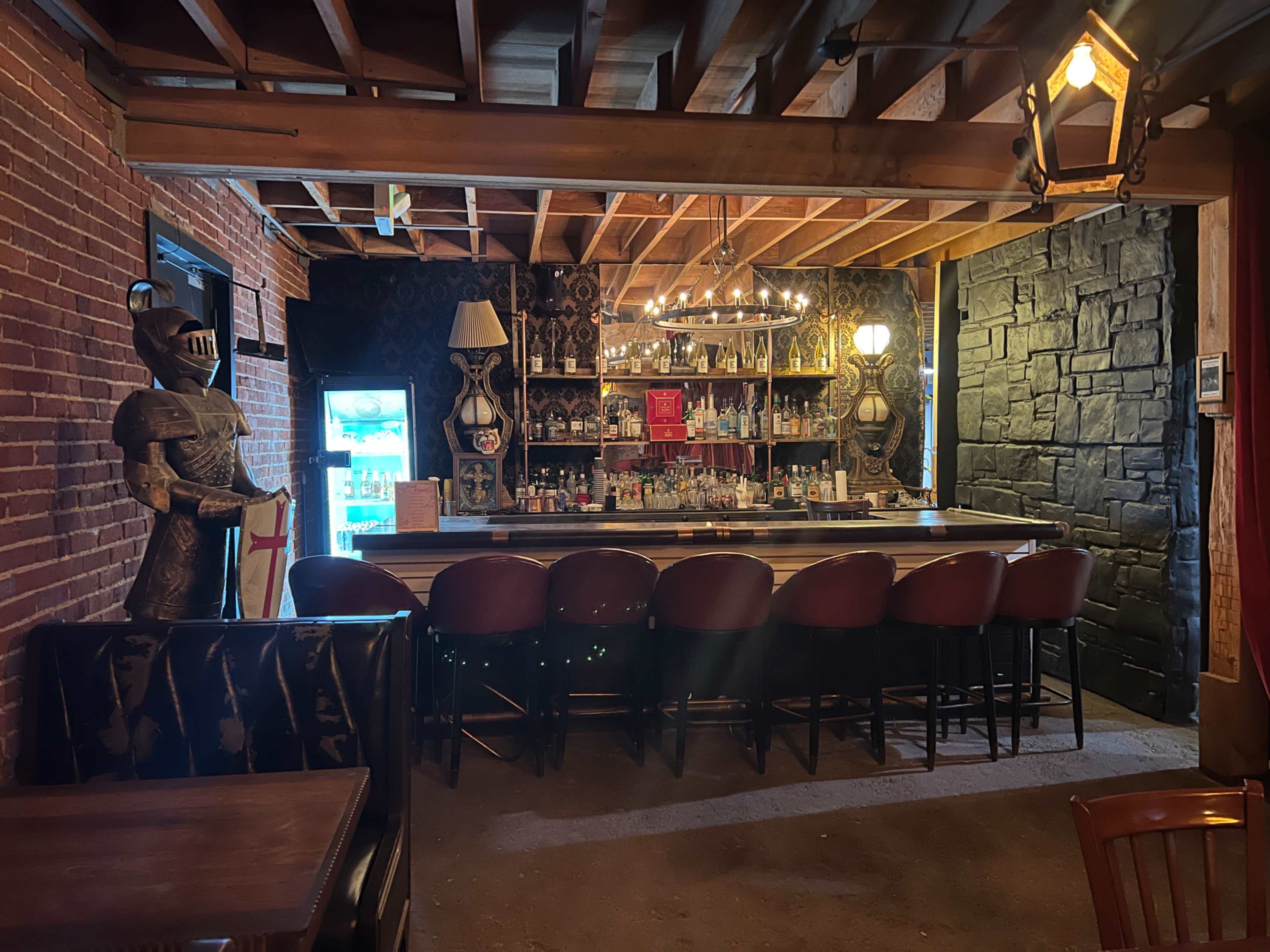 A bar area with a wooden counter, surrounded by high-backed chairs, and featuring medieval knight decorations and shelves lined with various bottles.
