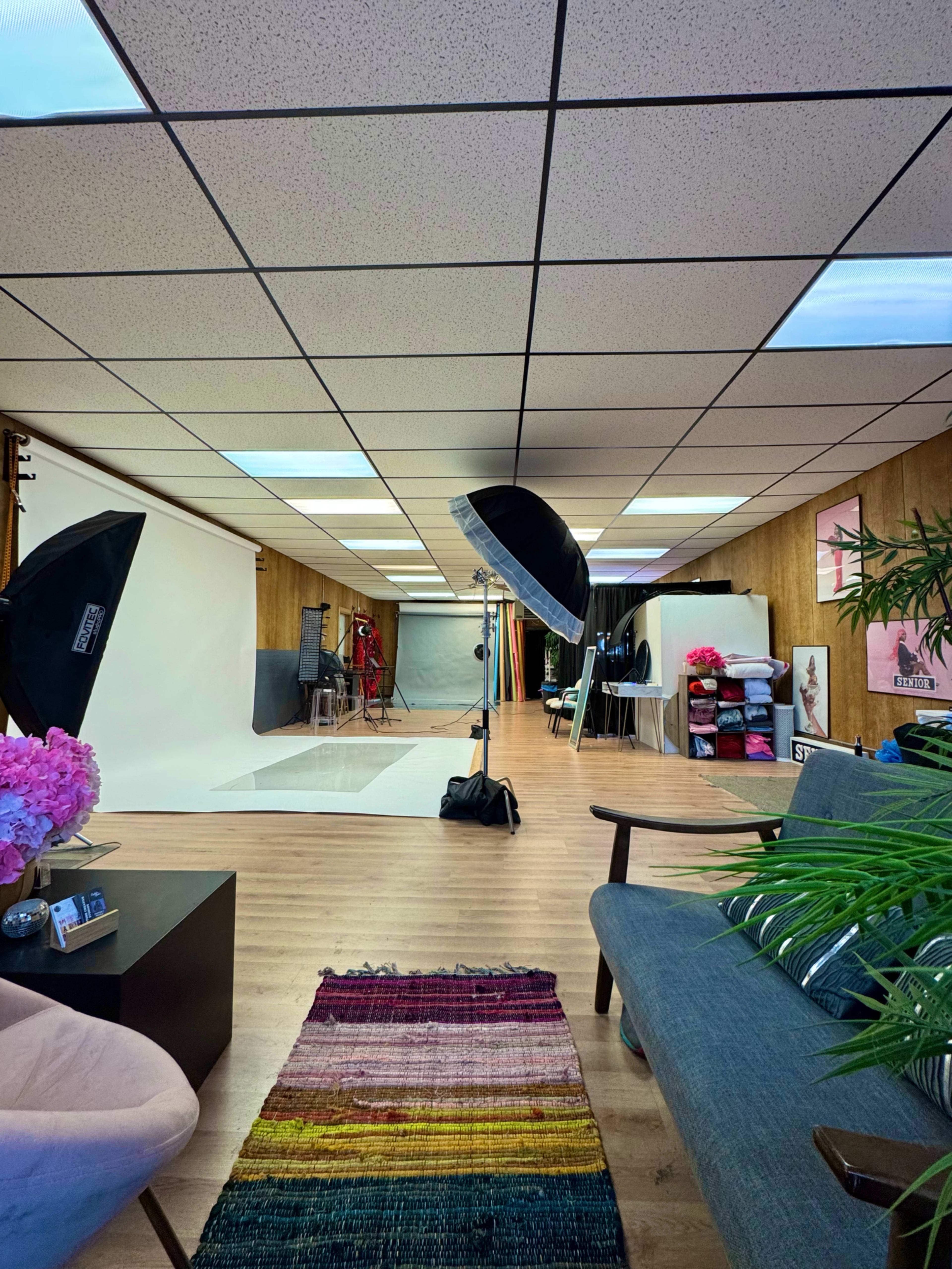 The image shows a photography studio with softbox lights, a backdrop area, and various props organized on shelves, along with a seating area featuring a rug and couch.