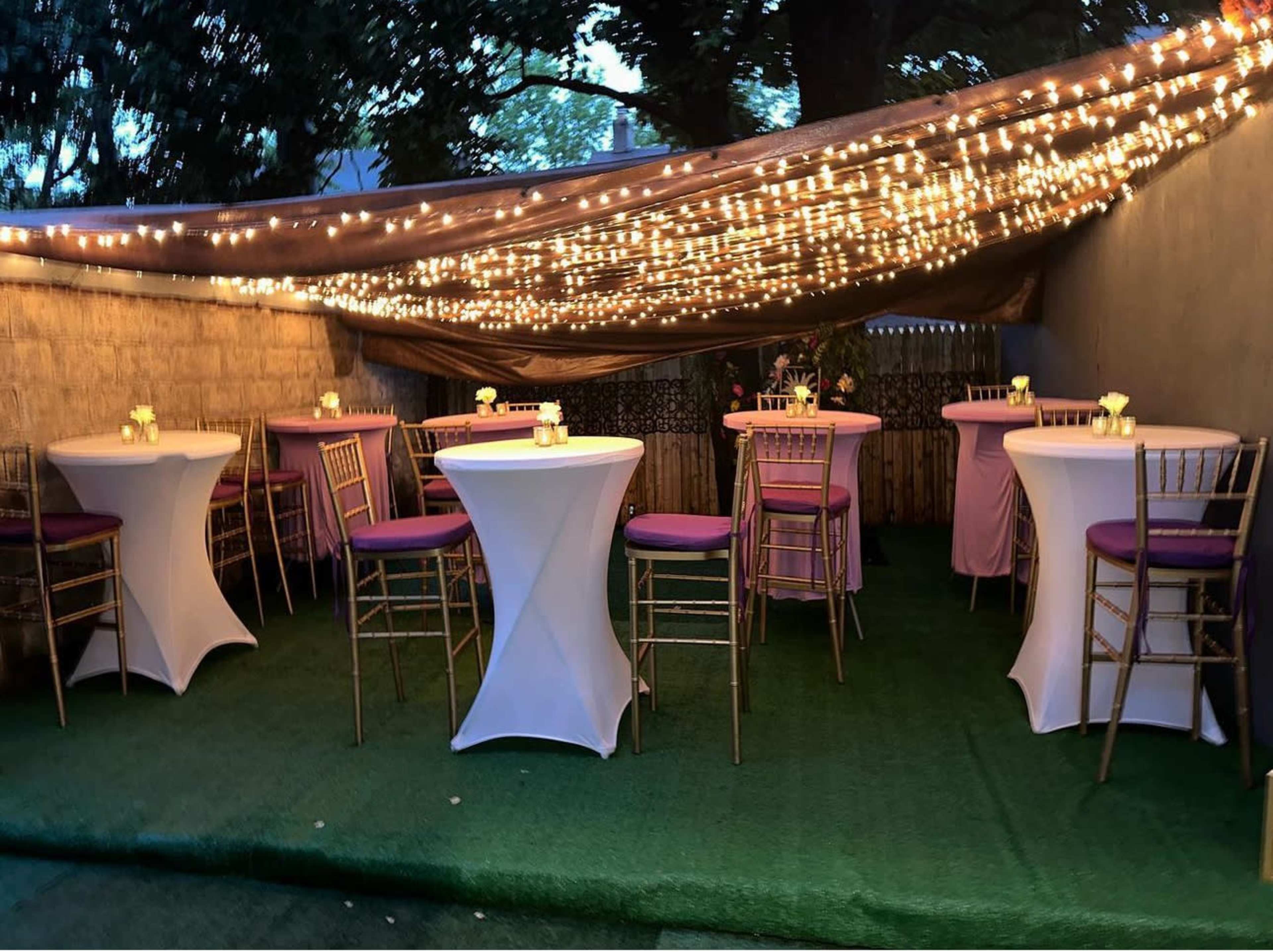 An outdoor party space with round tables draped in white and topped with purple cloth, under a canopy of string lights.