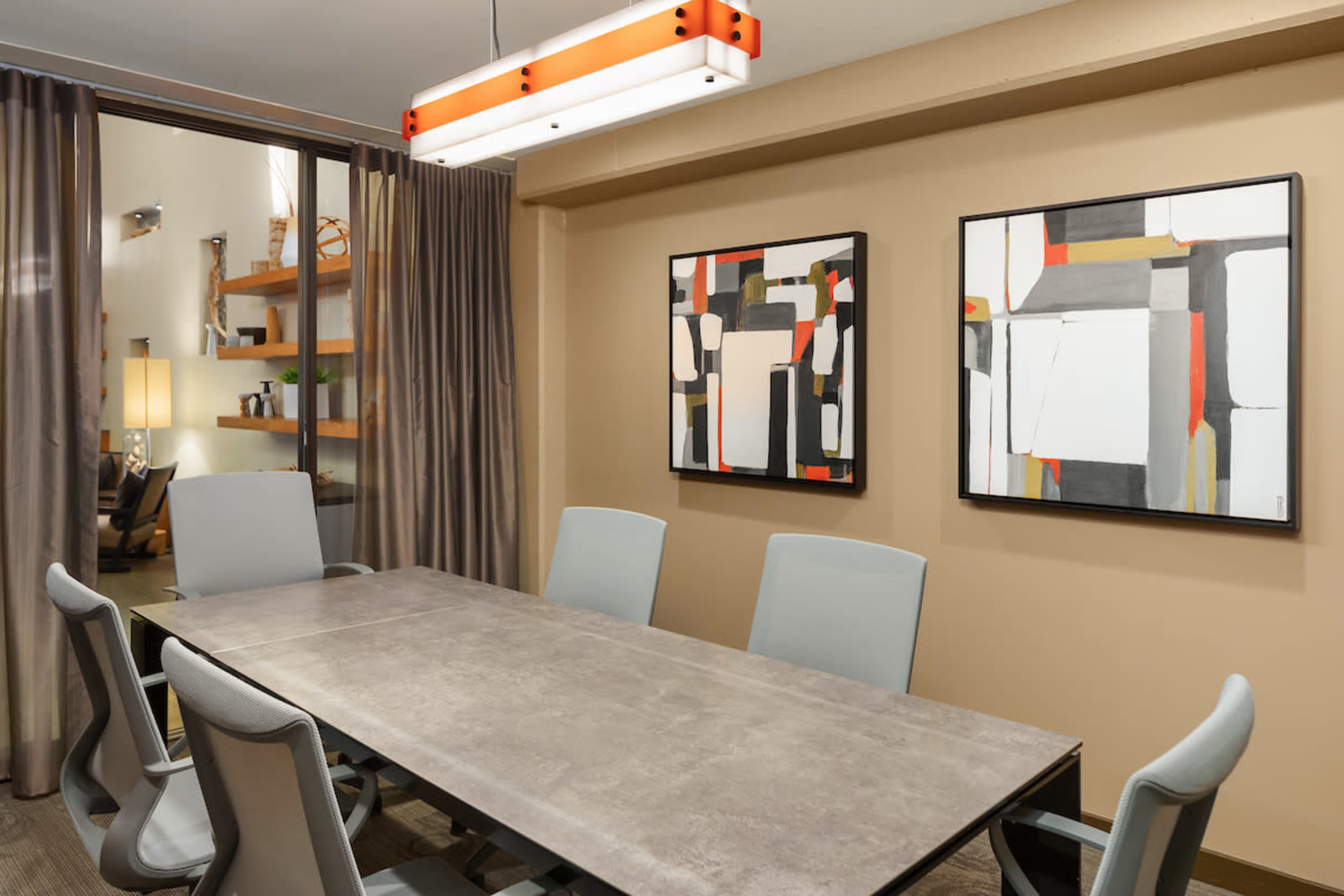 A modern conference room features a long table surrounded by light gray chairs, with two abstract paintings on the walls and a hanging light fixture overhead.