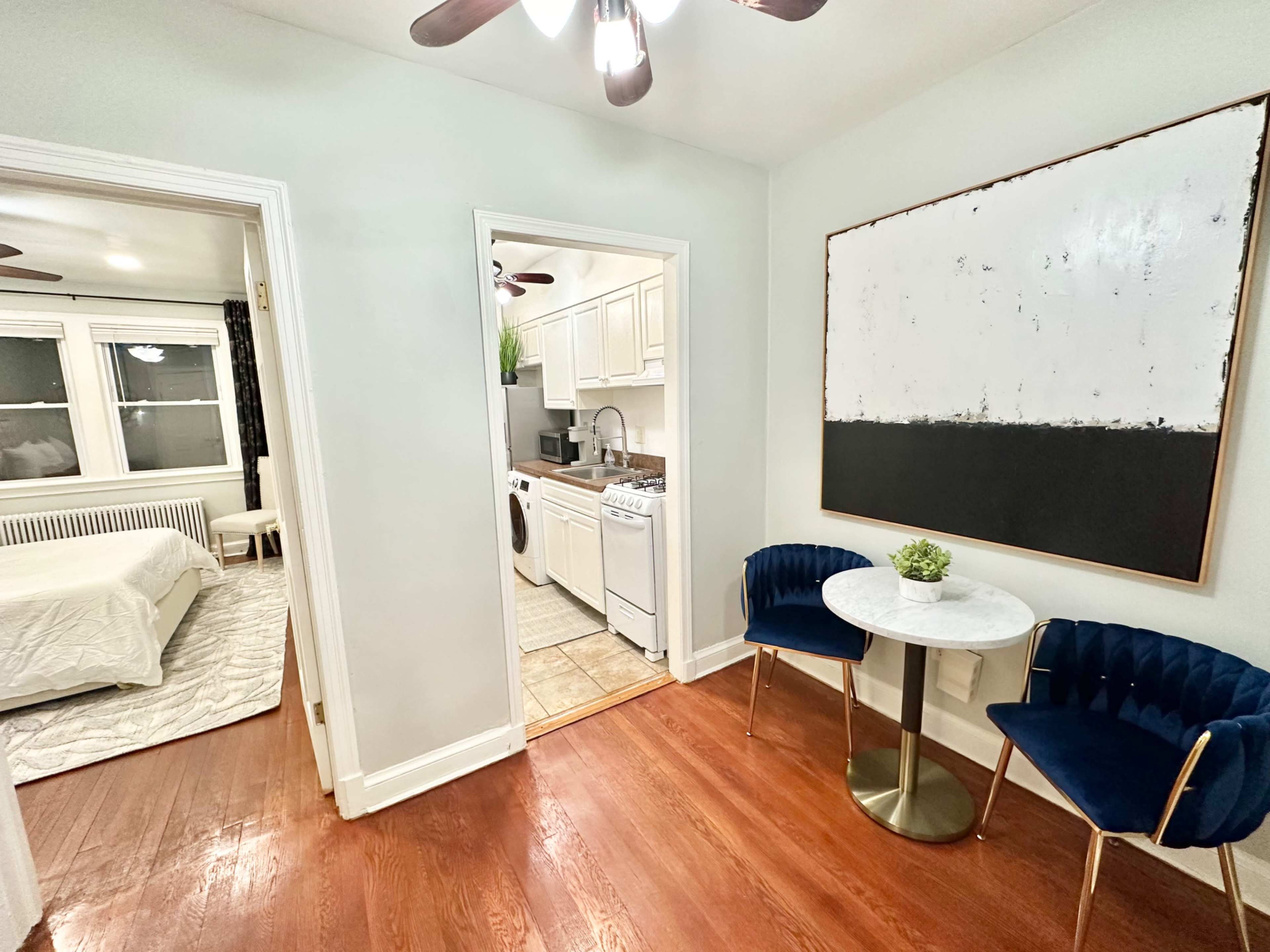 A small apartment hallway leading to a kitchen, connected to a bedroom, with a circular table and two chairs in the foreground.