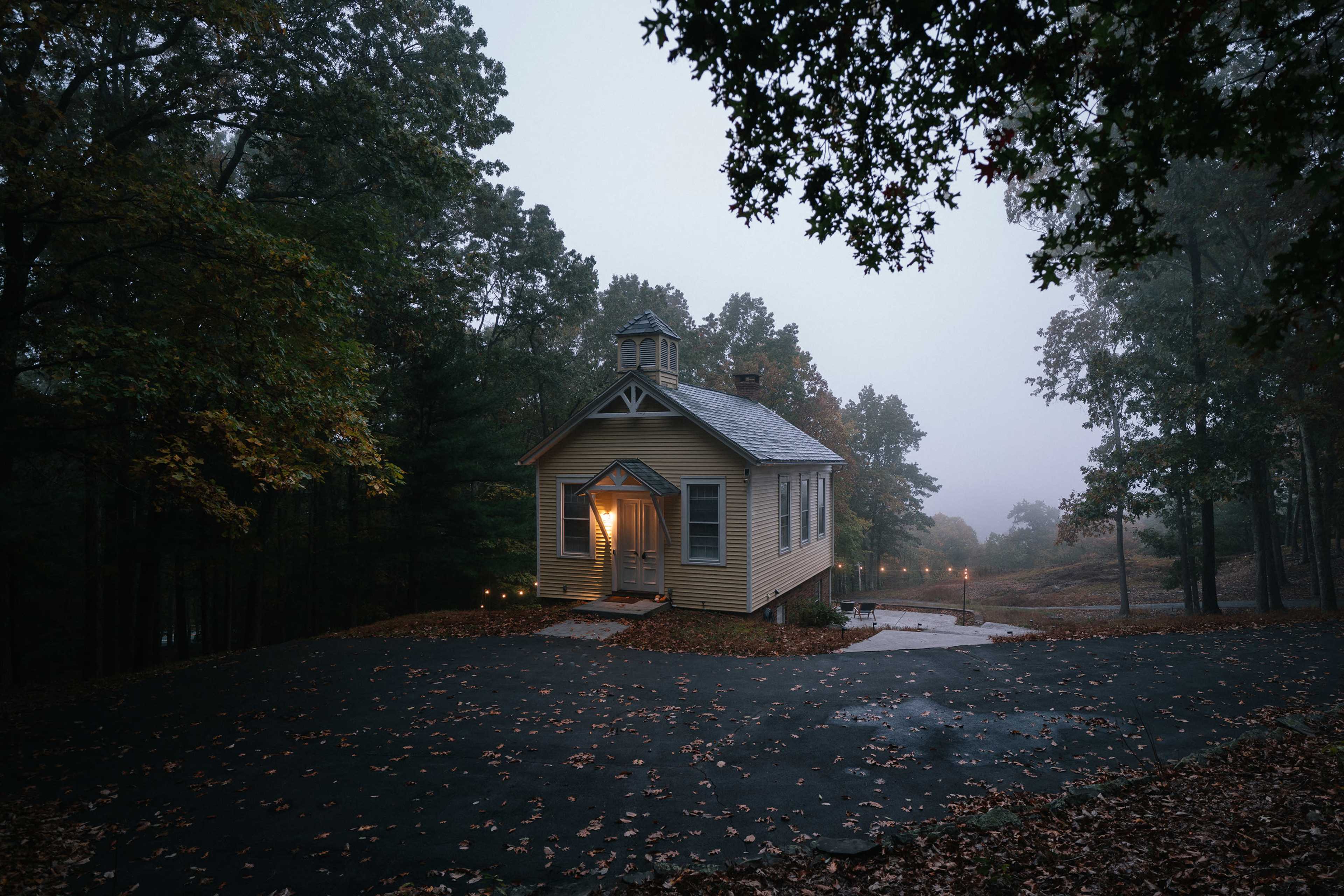A small, yellow house with a peaked roof sits on a winding driveway surrounded by trees in a foggy setting.