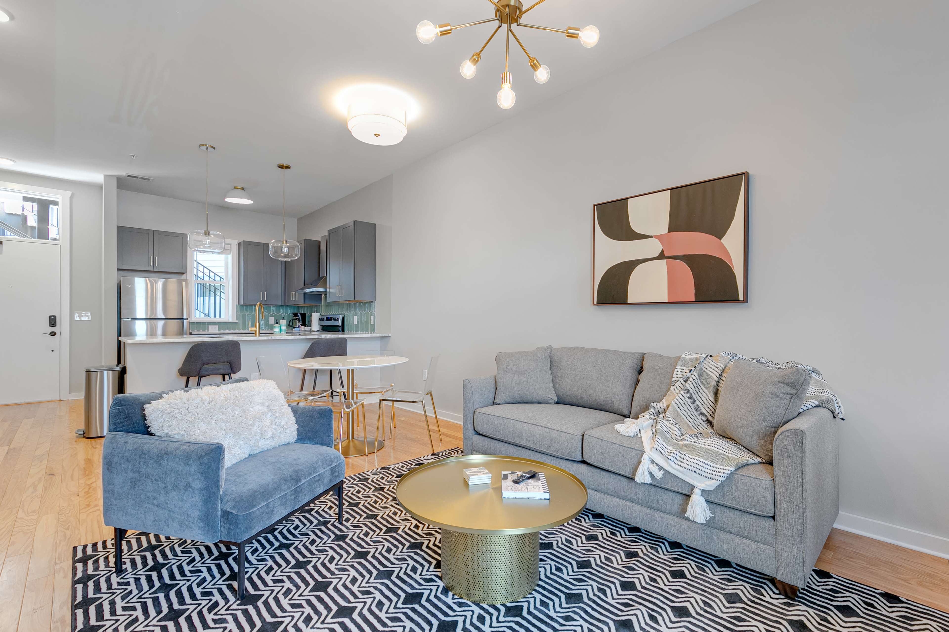 A modern living space featuring a gray couch, a blue accent chair, a round gold coffee table, and a dining area with a small white table and chairs.